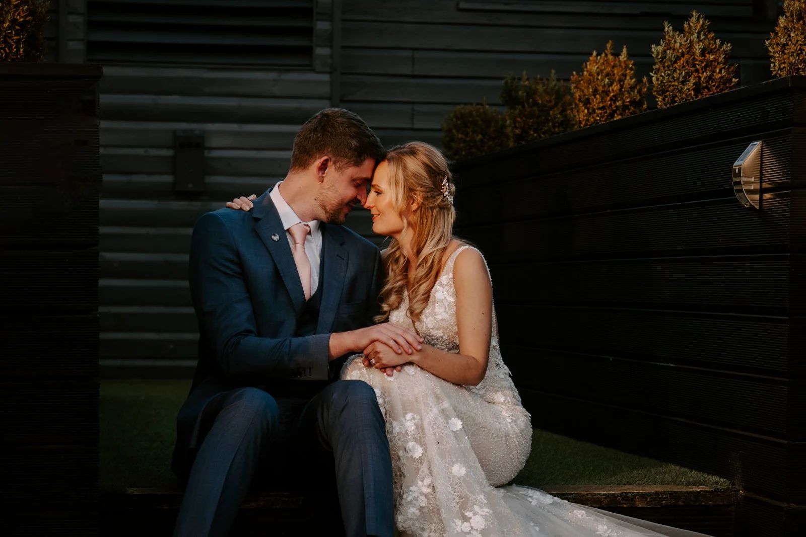 A bride and groom sitting close together with their foreheads touching and eyes closed, outdoors at sunset, against a dark wooden fence background.