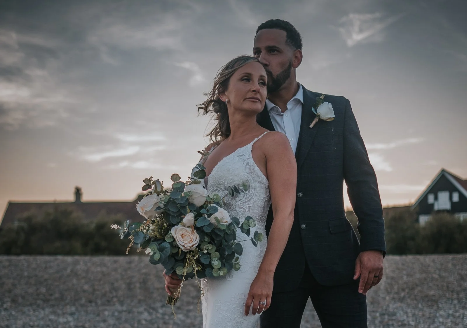 A bride and groom standing outdoors at sunset, with the bride holding a bouquet of flowers, both dressed in wedding attire, and a cloudy sky overhead.