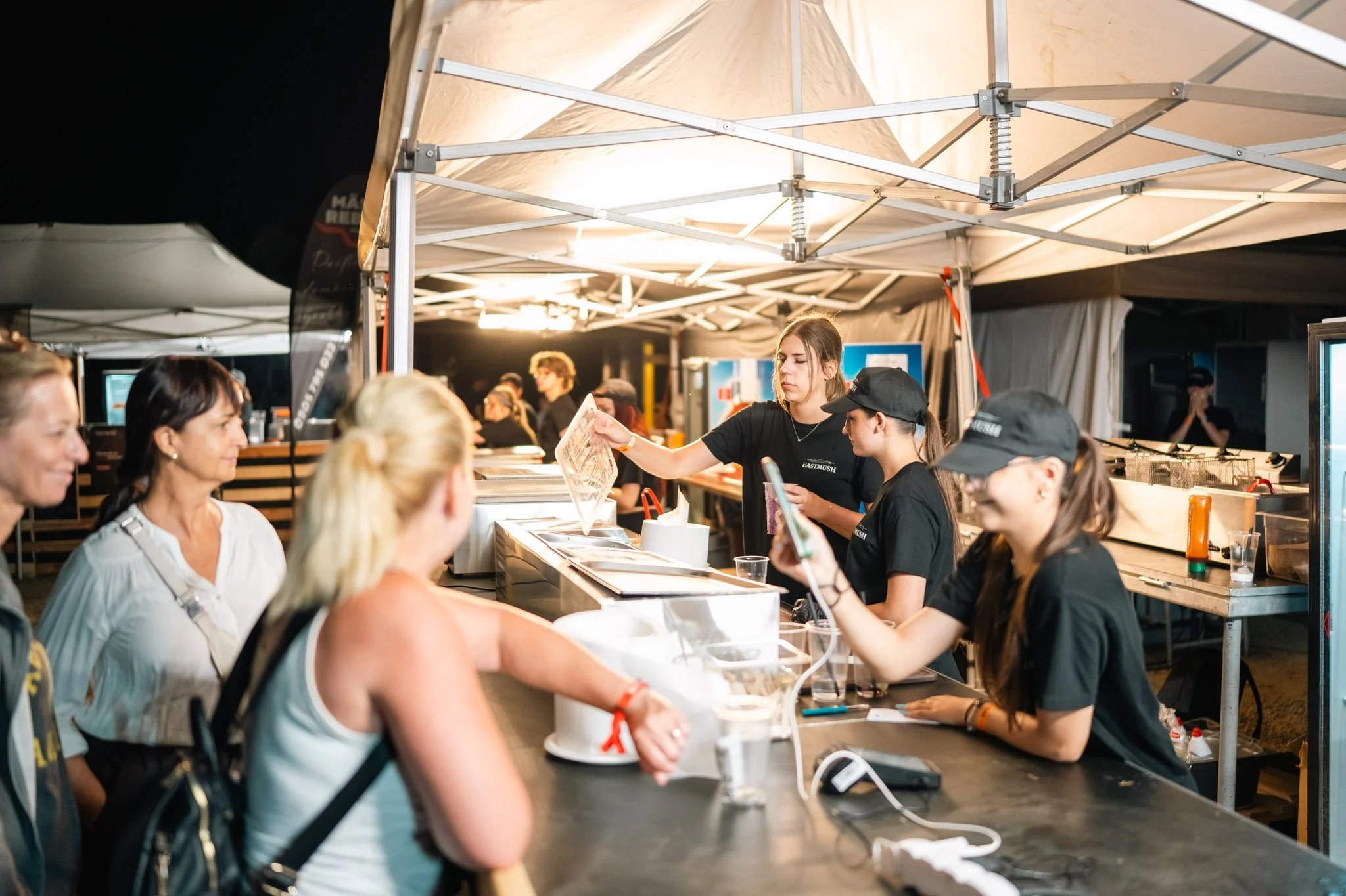 People lining up at a food or drink stand at night, with a canopy overhead and bright lighting illuminating the scene.