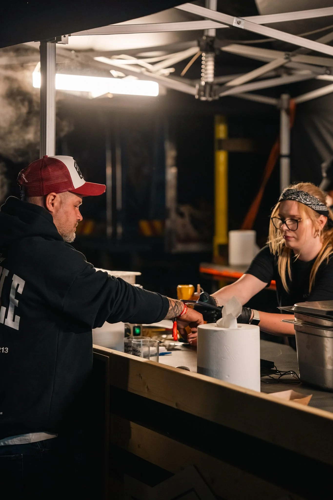 Two people working at a food or drink stall under a black and white canopy, with a roll of paper towels and various supplies on the counter.