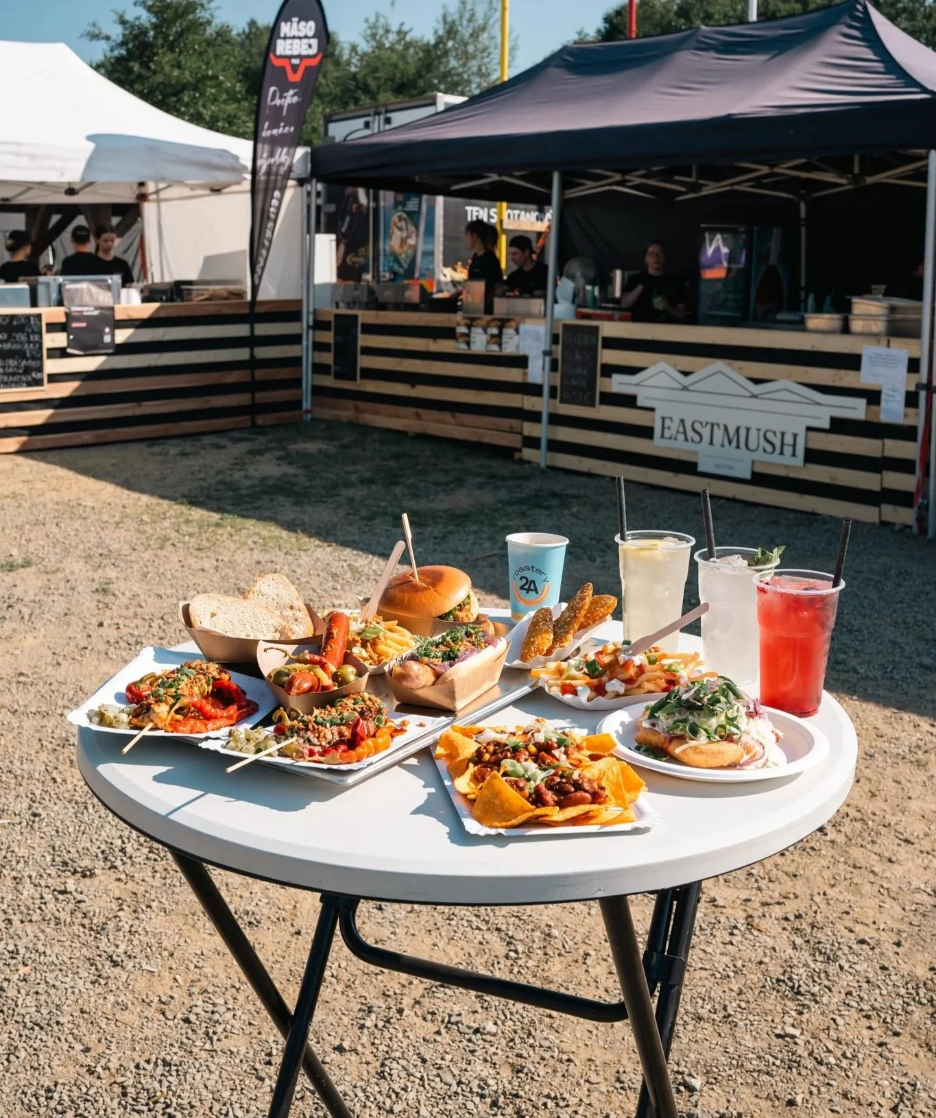 Outdoor food festival with a white table holding various dishes like tacos, nachos, sandwiches, and drinks, with food stalls in the background.