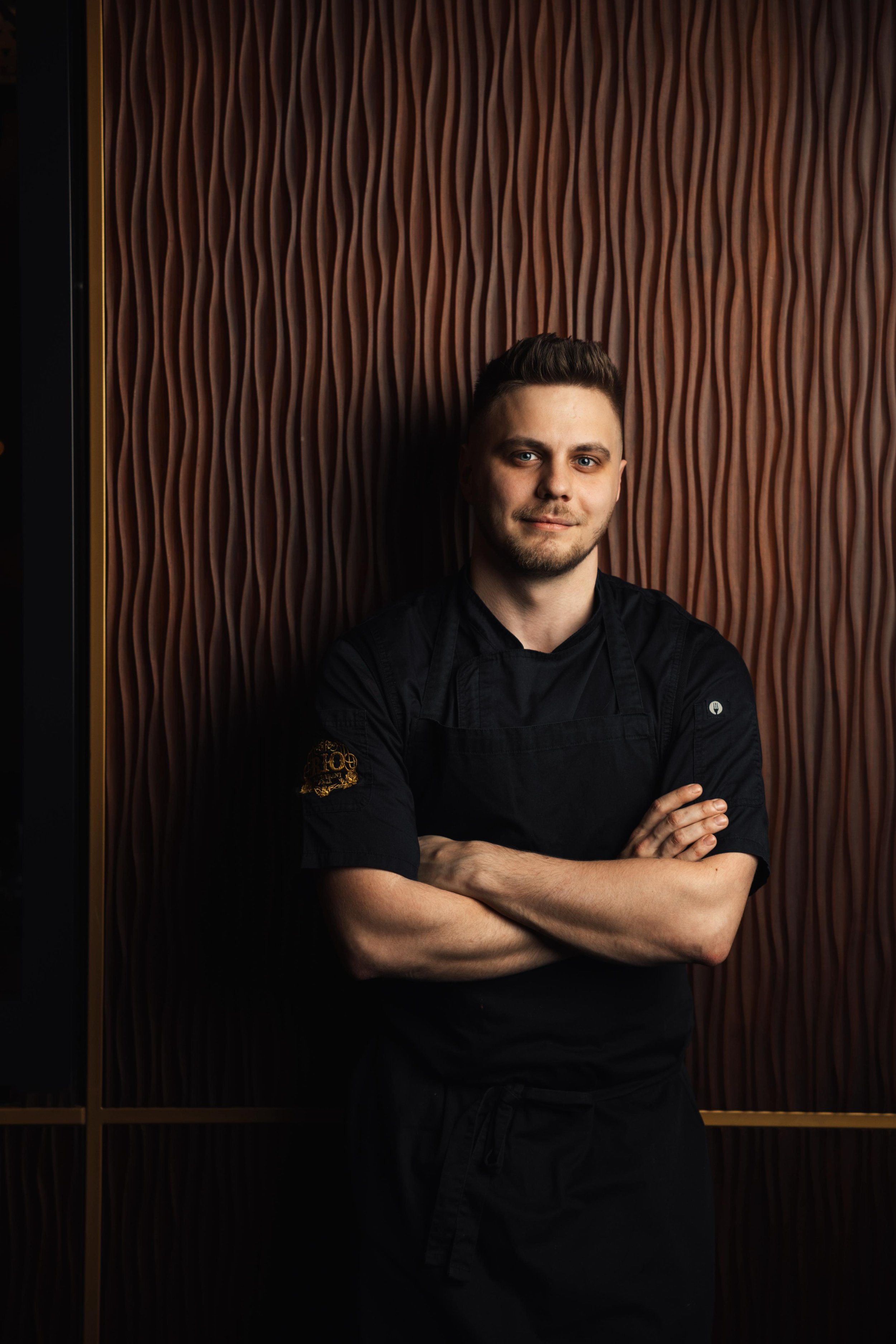 Portrait of a young male chef with arms crossed, standing in front of a textured wooden wall.