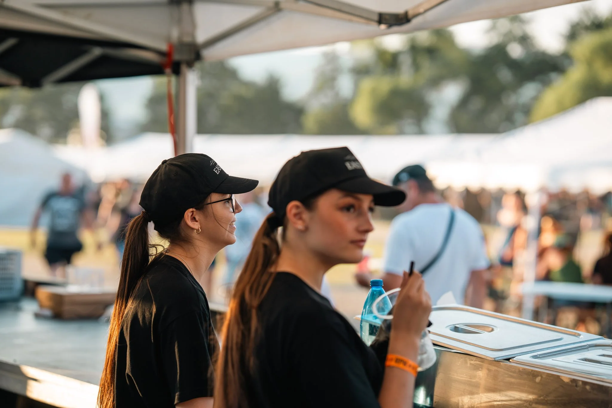 Two women with dark hair wearing black caps, standing in line at an outdoor food or drink stand. One woman is smiling, while the other is holding a water bottle and looking forward. There are other people in the background, some wearing hats, and a canopy overhead with a blurry outdoor scene of trees and bright daylight.