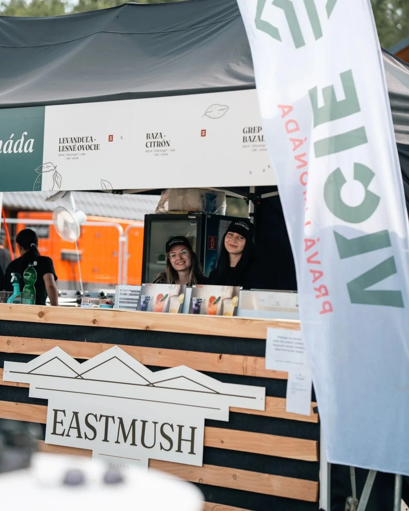 Two smiling women working at an outdoor food stand labeled 'EASTMUSH', with menu items and colorful drinks displayed in front.