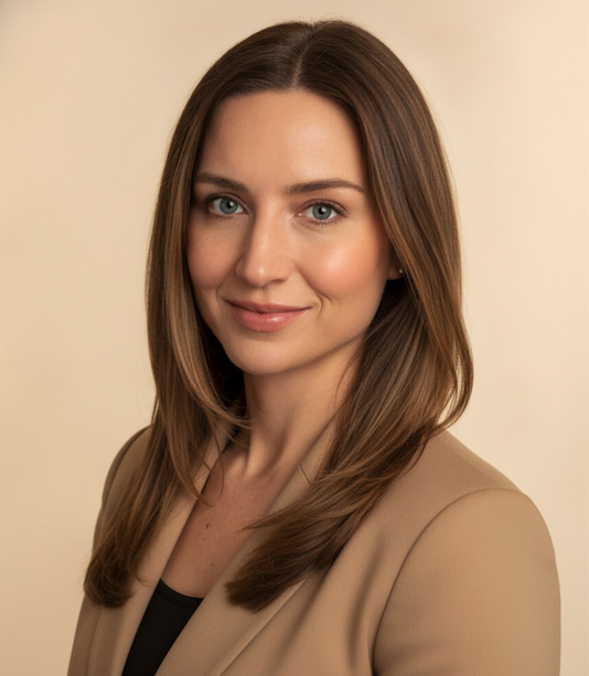 A young woman with long, light brown hair, blue eyes, and fair skin, wearing a beige blazer over a black top, smiling softly against a neutral background.