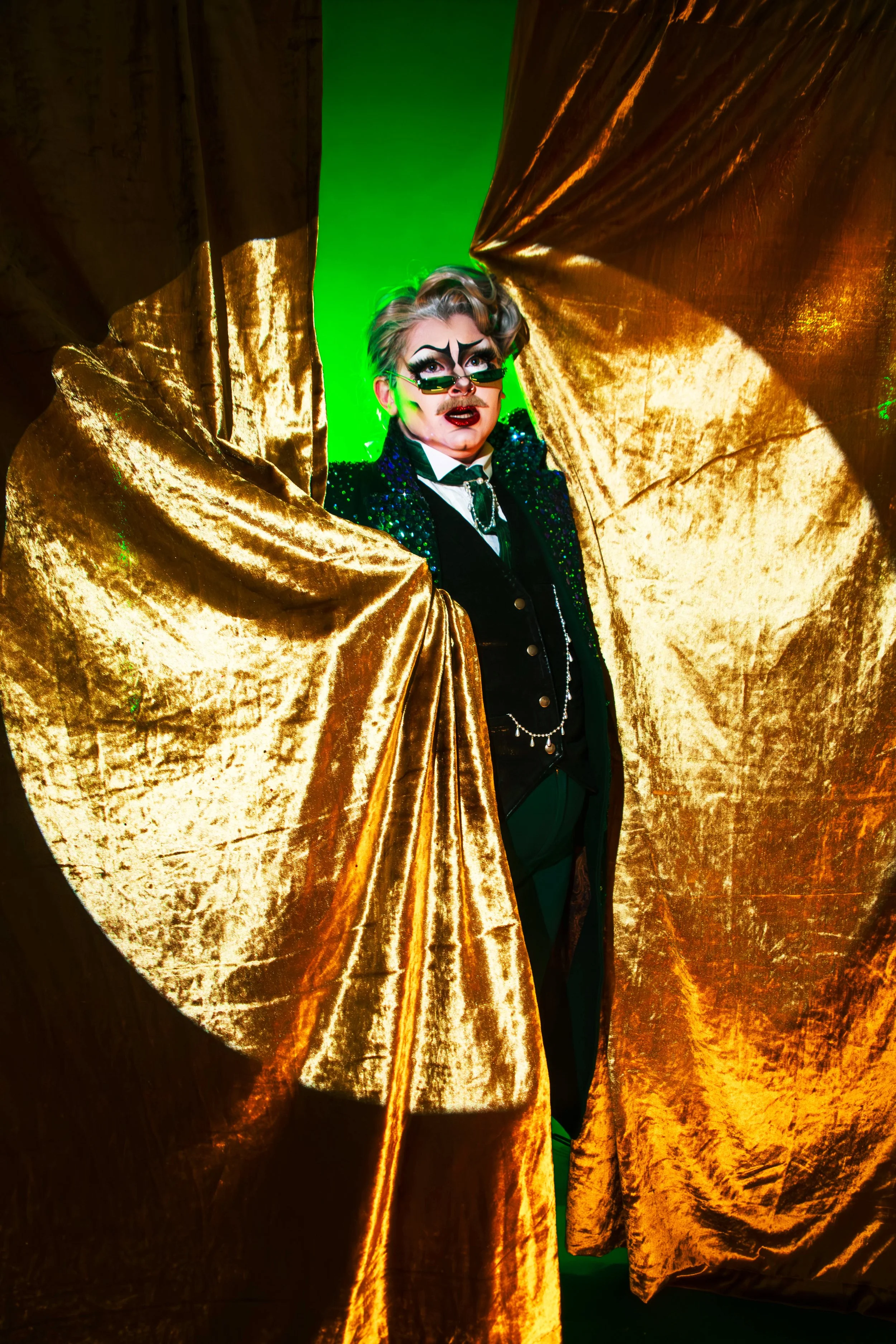 A person dressed in a theatrical costume with dramatic makeup, wearing small, green-tinted sunglasses, peeking through gold velvet curtains with a green background.