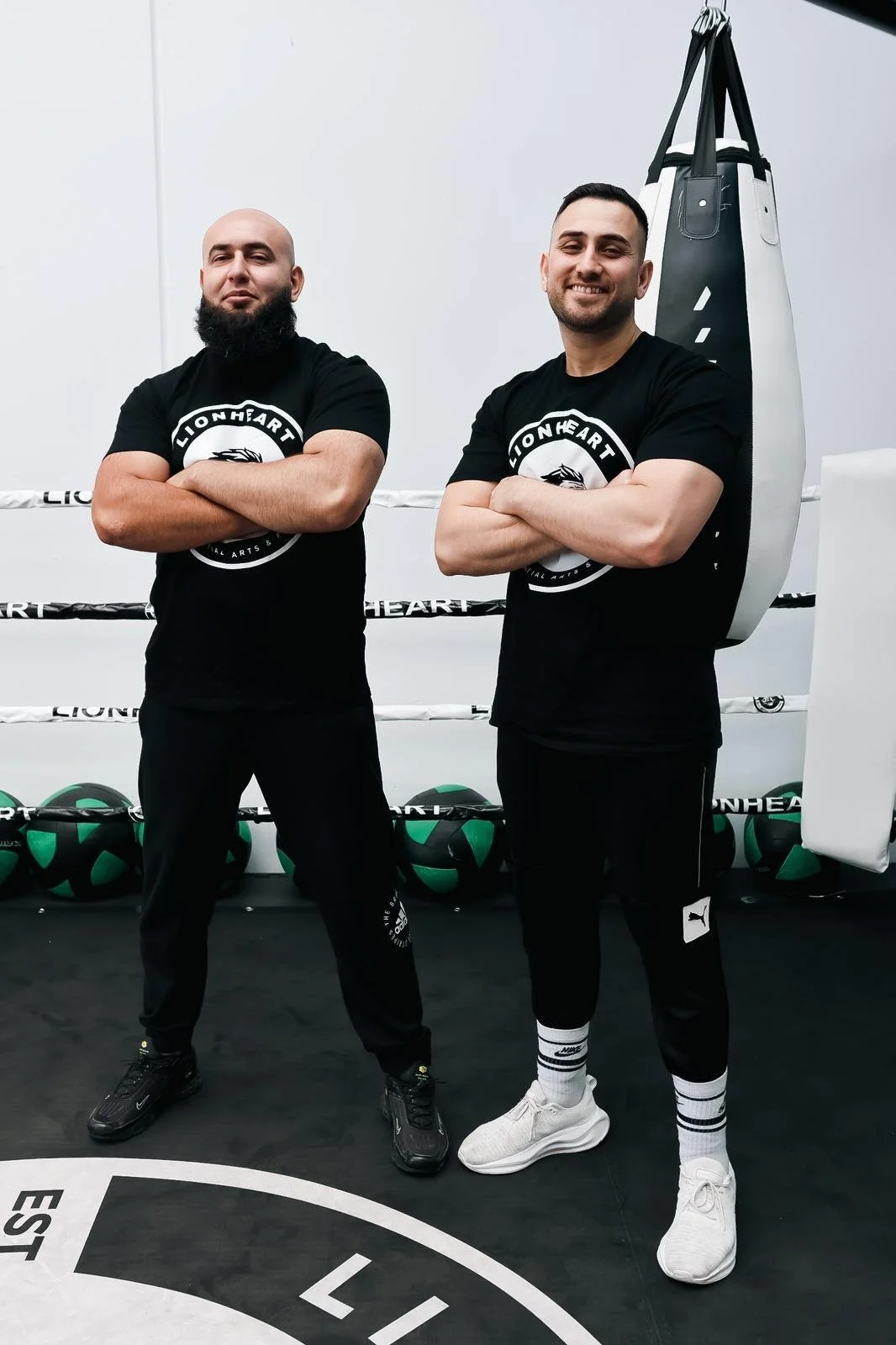 Two men standing with arms crossed in a boxing gym, wearing black T-shirts with 'LIONHEART' logo, and black sweatpants. The gym has boxing gloves and a punching bag visible in the background.