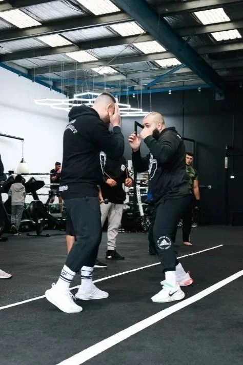 Two men are sparring in a gym, wearing black hoodies and white sneakers, with one man holding up his hands in a defensive stance and the other raising his fists. Several people are in the background, and the gym has a black floor and modern ceiling lighting.