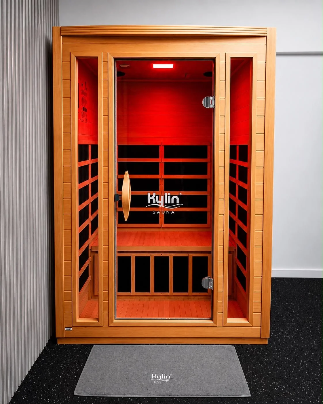 A wooden infrared sauna with a glass door, inside red lighting, and a small wooden bench. The brand name 'Kylin Sauna' is visible on the door and the mat outside.