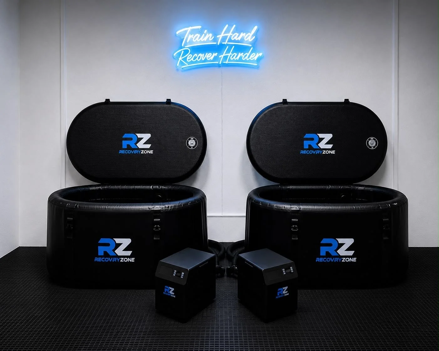Two black recovery tanks labeled 'RZ Recovery Zone' with their lids open, placed side by side on a black textured floor. Two black boxes with digital displays and labeled 'RZ' are in front of the tanks. A neon sign on the white wall behind reads 'Train Hard, Recover Harder' in blue cursive.