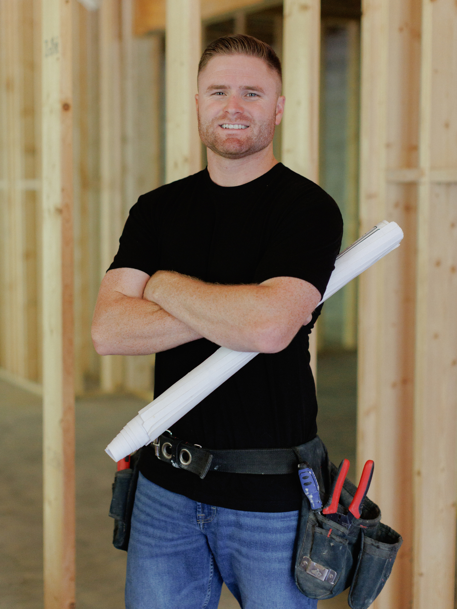 Construction worker in a black t-shirt and blue jeans holding blueprints at a building site with wooden framing in the background.