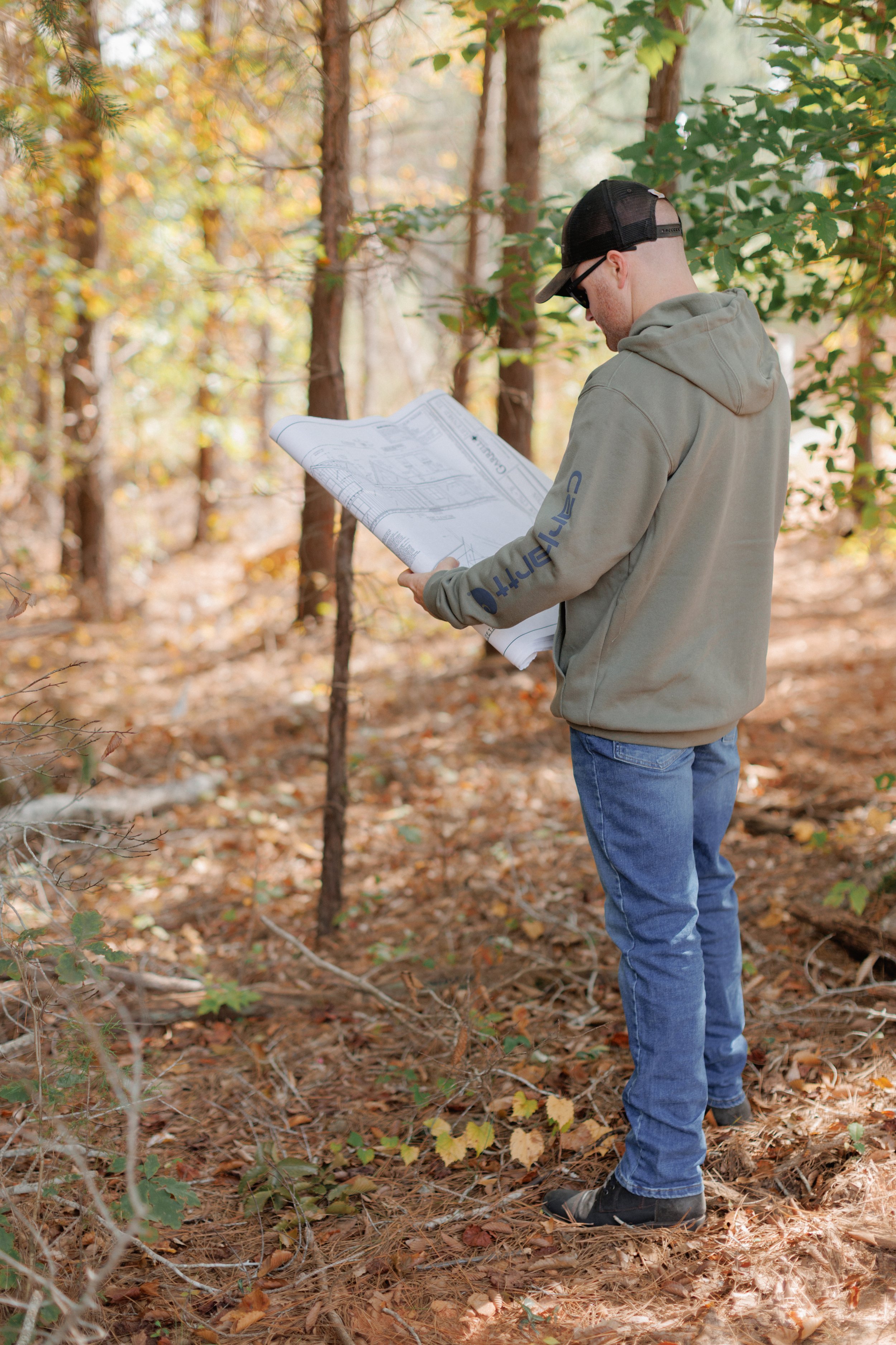 A man in a green hoodie, blue jeans, and black shoes is standing in a forest, reading a blueprint or map. He is wearing sunglasses and a black trucker hat, and the forest is filled with trees and fallen leaves.