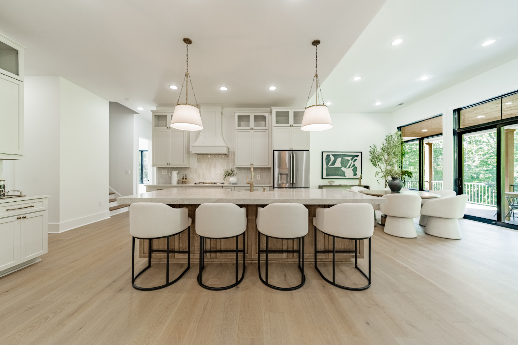 Open-concept kitchen and living space with white cabinetry, a large island with four barstools, and a sitting area with white chairs and large windows showing a green outdoor view.