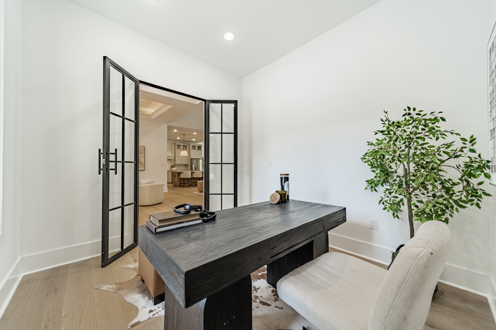 Home office with black desk, beige chair, potted plant, and decorative items, view into living area with kitchen in the background.