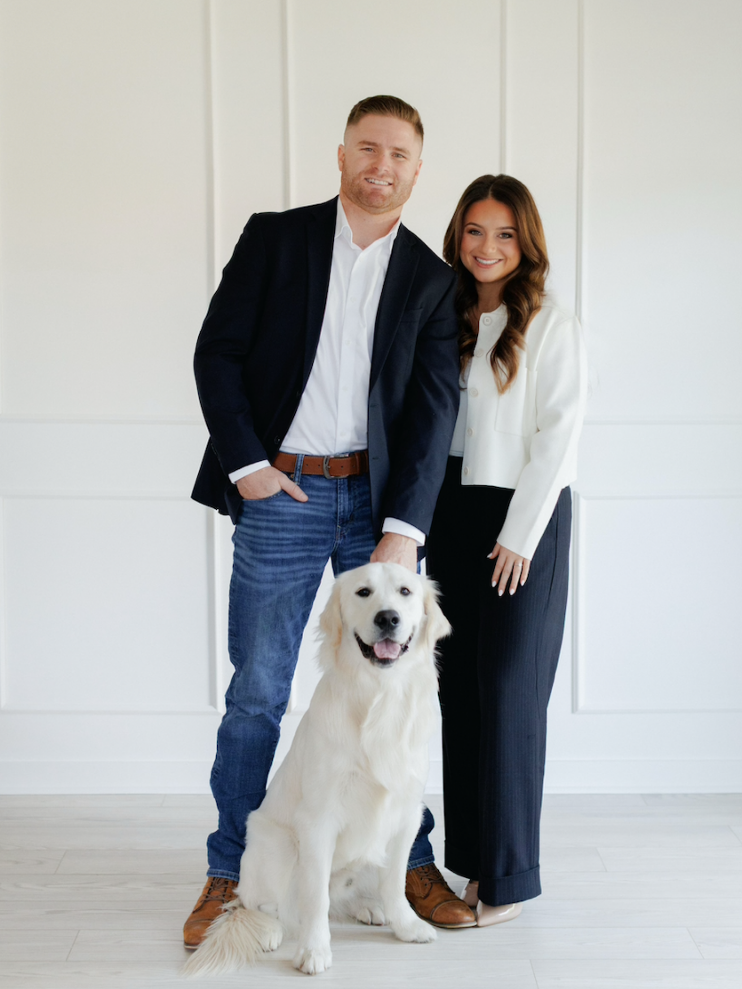 A man and a woman standing with a golden retriever dog in front of a white wall. The man is wearing a navy blazer over a white shirt and jeans, and the woman is dressed in a white blazer and black trousers.