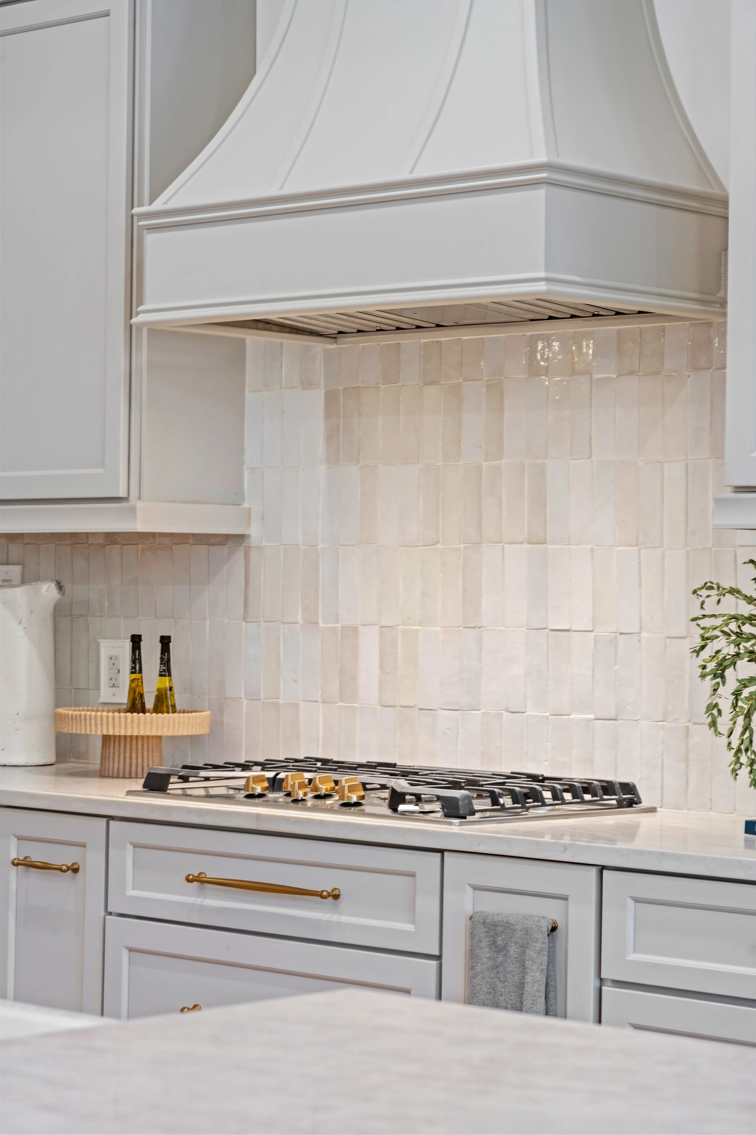 White kitchen with cabinets, a stove, and a white tile backsplash. There is a round wooden tray with bottles on the countertop and a towel hanging from one of the cabinet handles.