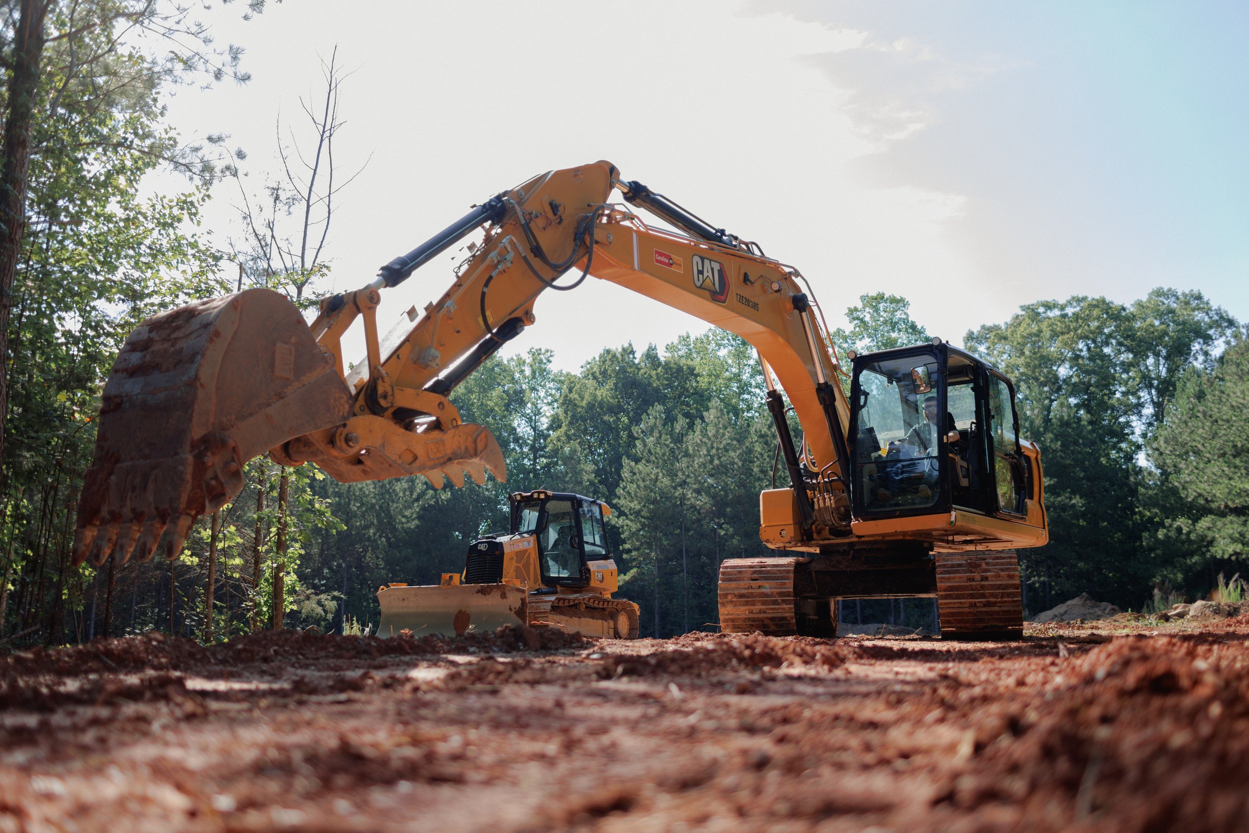 A construction excavator with a large bucket attachment working on a dirt site surrounded by trees.