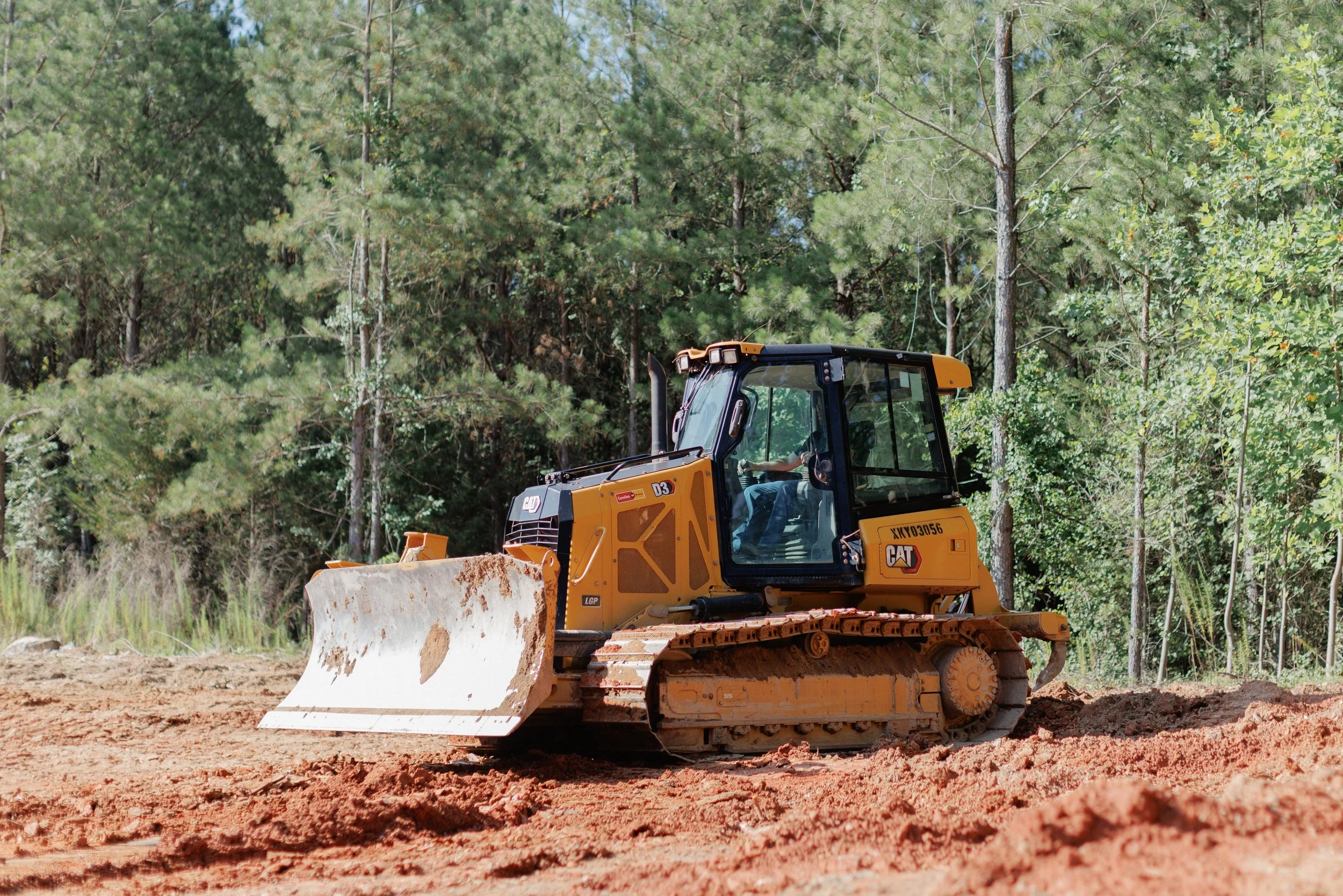 A yellow Caterpillar bulldozer working on a dirt construction site surrounded by green trees.