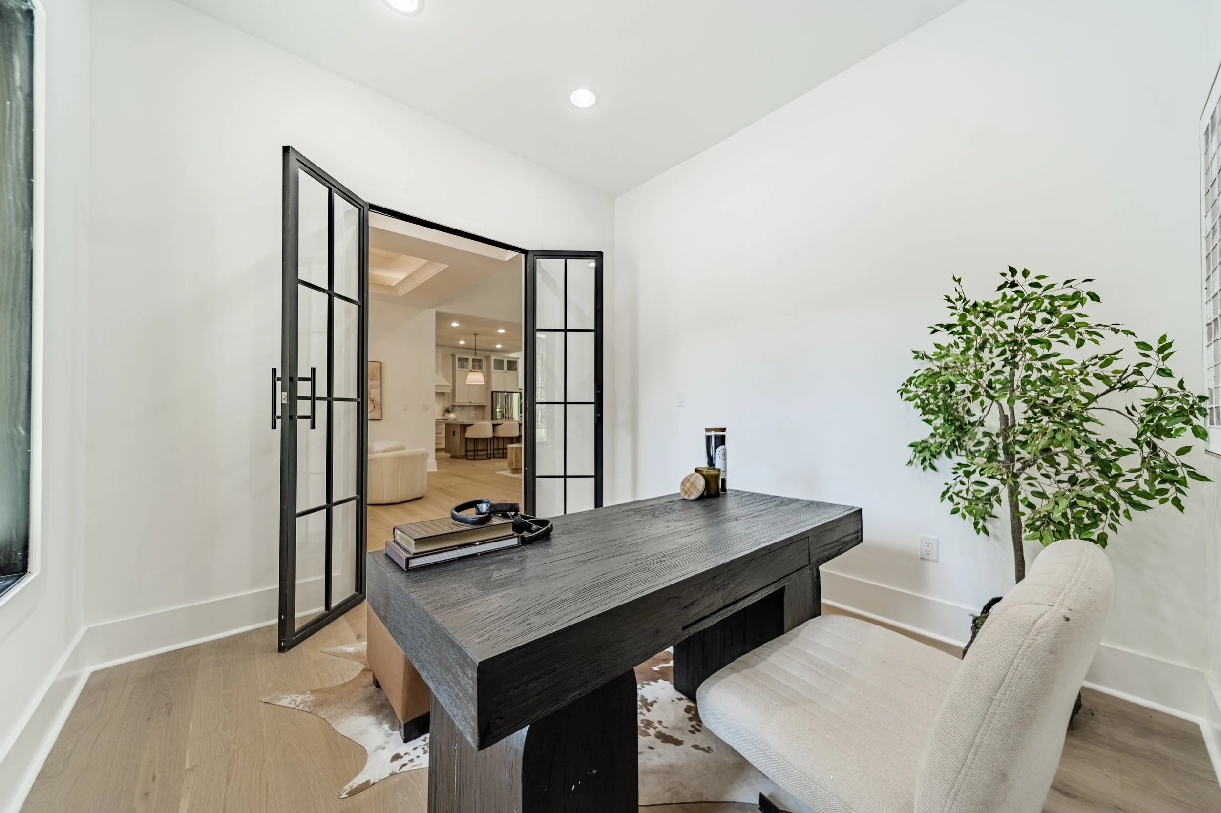 Modern living room with white walls, black-framed glass double doors, a wooden table with books and decor, a beige couch, a tall green plant, and a cowhide rug, with an open view into a kitchen and dining area.