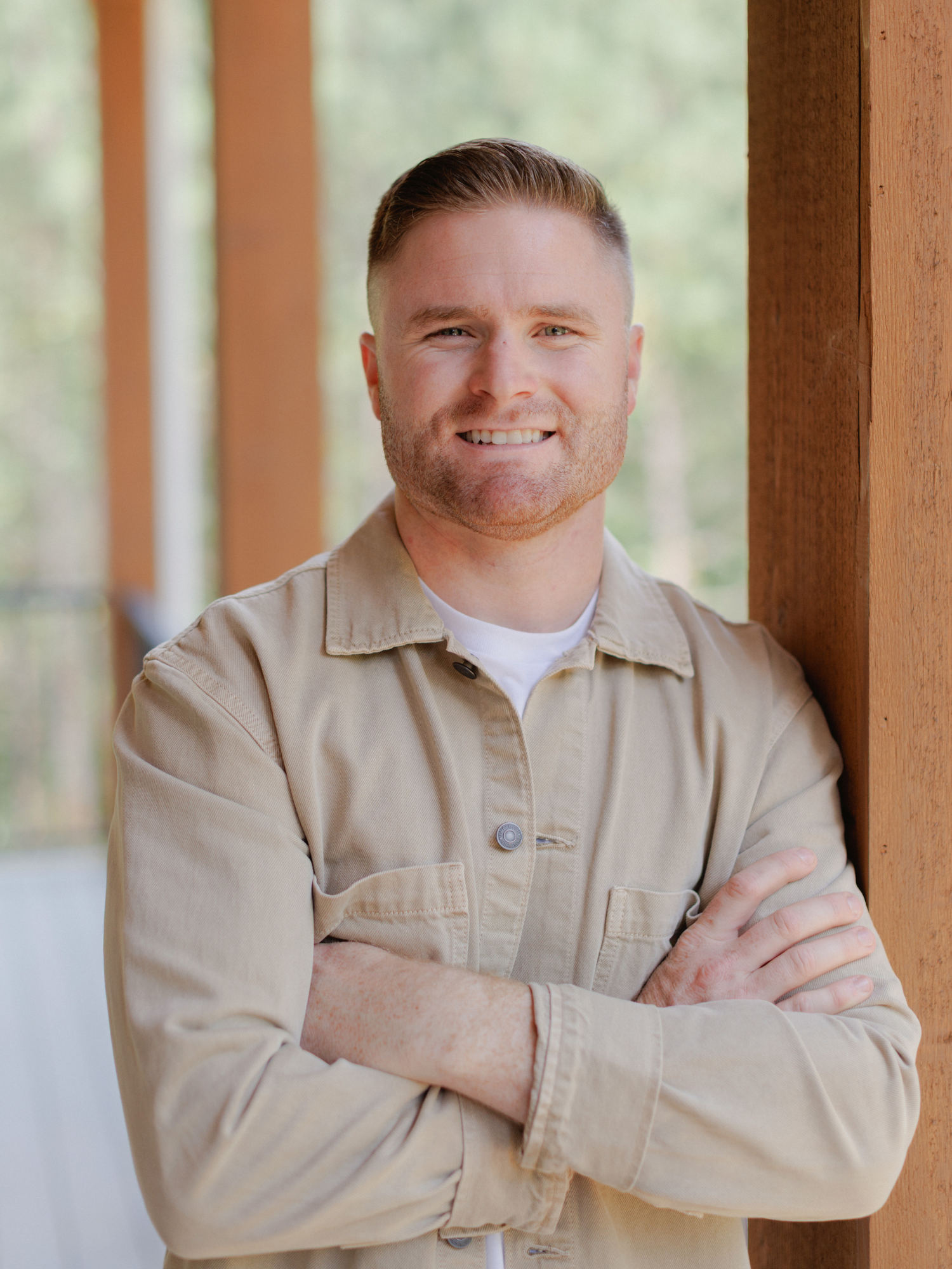 A young man with short hair and a beard, smiling, standing with arms crossed in front of a wooden post, indoors with a blurred nature background.
