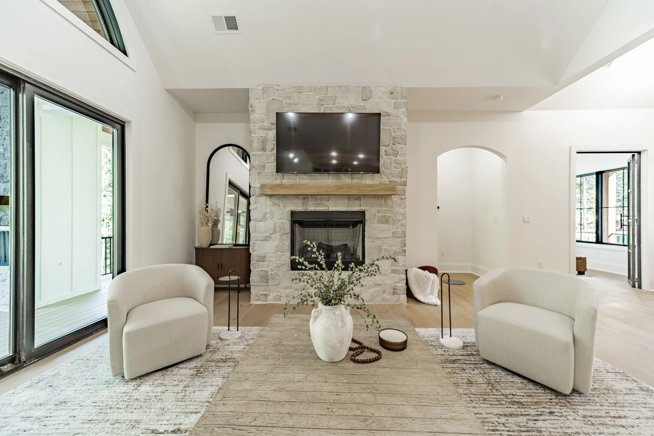 Living room with white walls, a stone fireplace, wall-mounted TV, beige armchairs, vases, mirror, and wooden accents, large glass sliding doors.