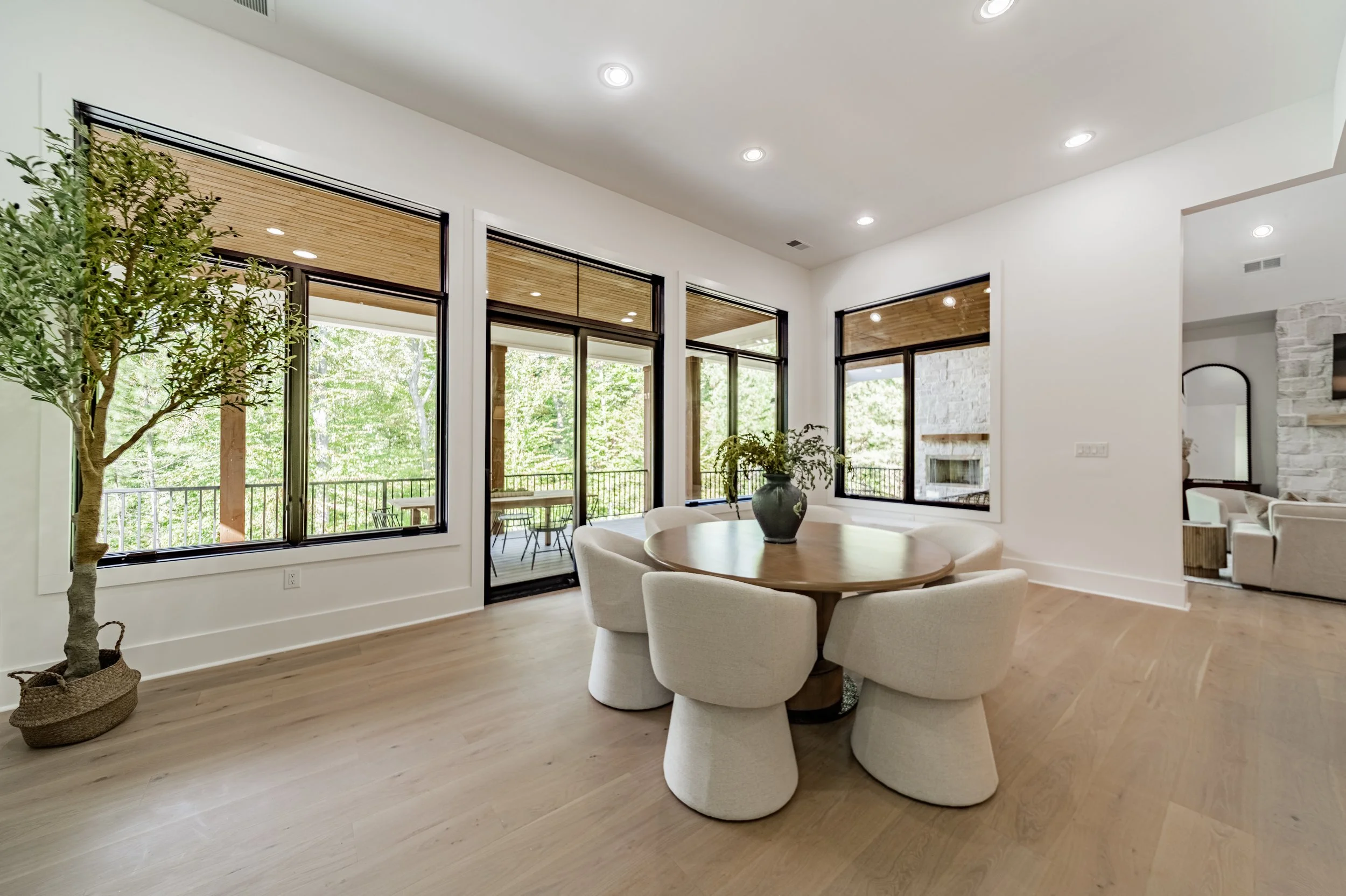 Bright living room with large windows, a round dining table with six white upholstered chairs, a potted tree in a woven basket, and a view of a balcony with outdoor seating surrounded by greenery.