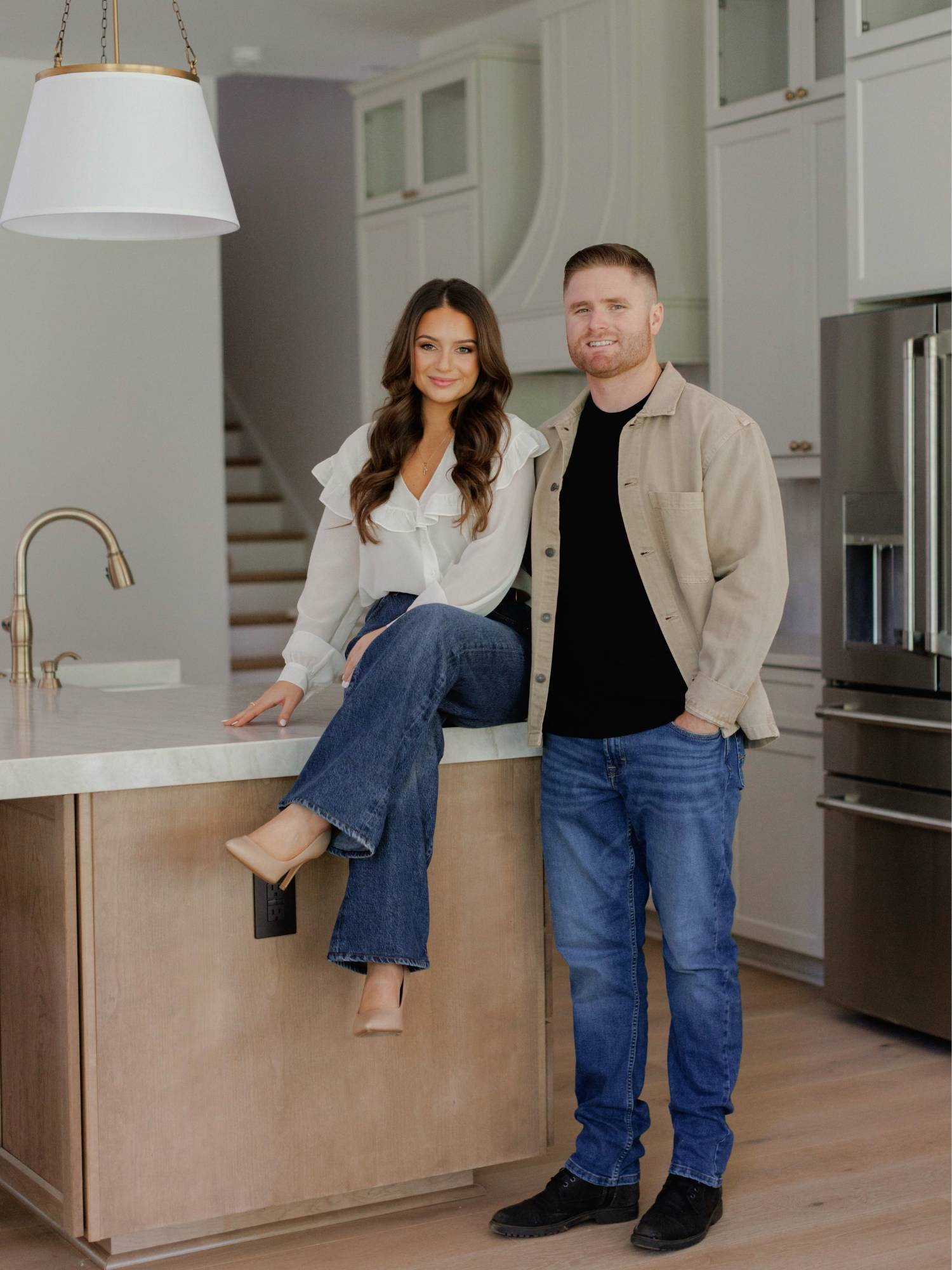 A man and woman standing in a modern kitchen, with the woman sitting on a kitchen island and the man standing beside her, both smiling at the camera.
