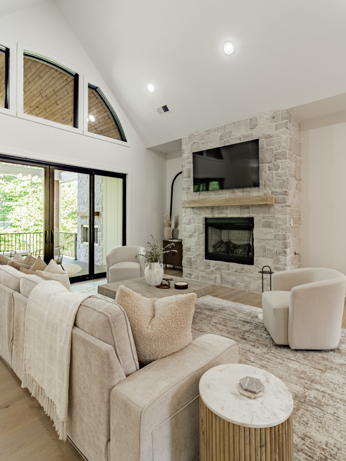 Living room with beige sofa, white armchairs, stone fireplace, flat-screen TV, and large windows with a view of a balcony and trees outside.