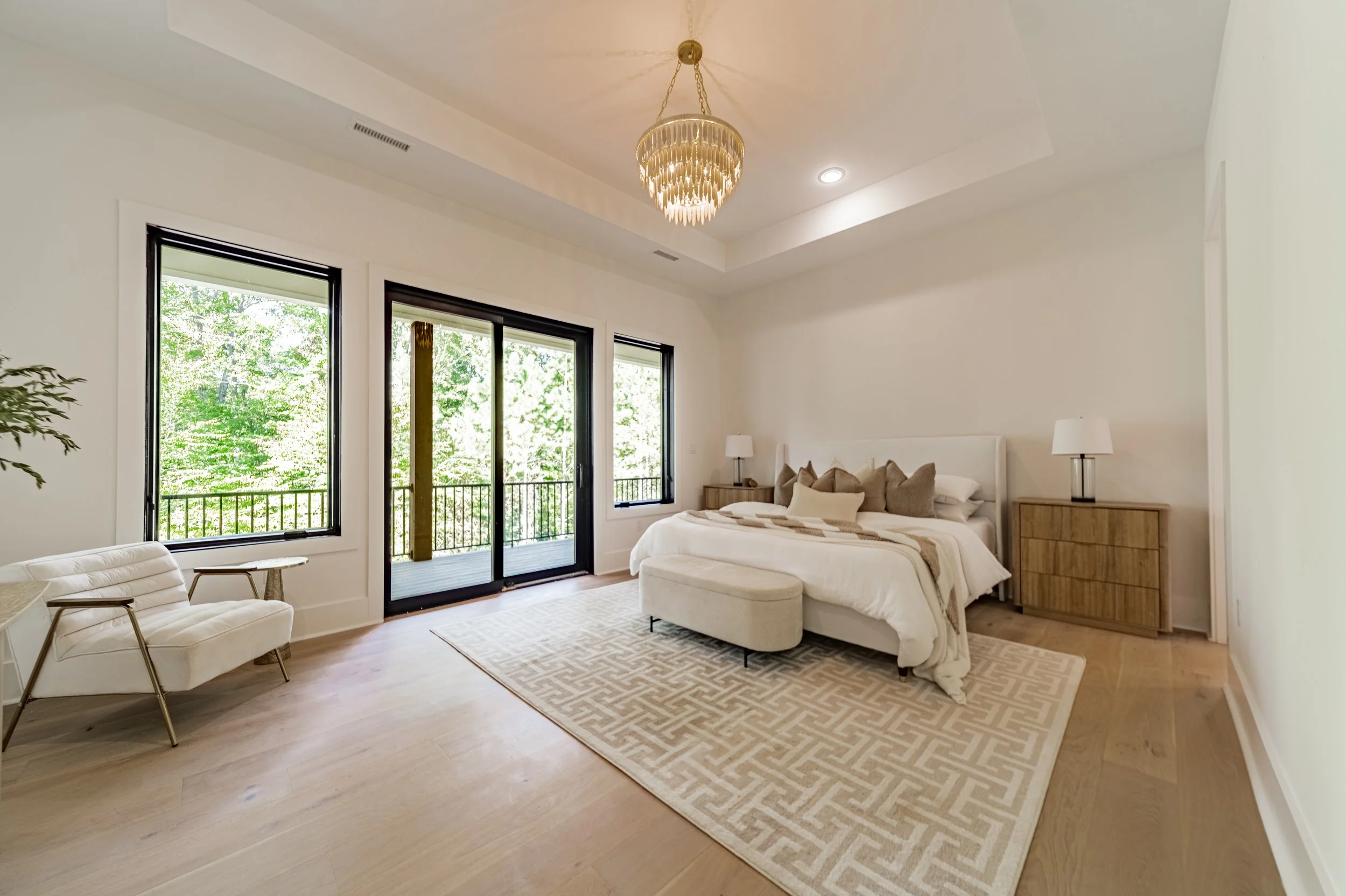 Bedroom with large windows, a bed with beige pillows, two wooden nightstands with lamps, a chaise lounge, an area rug, and a chandelier