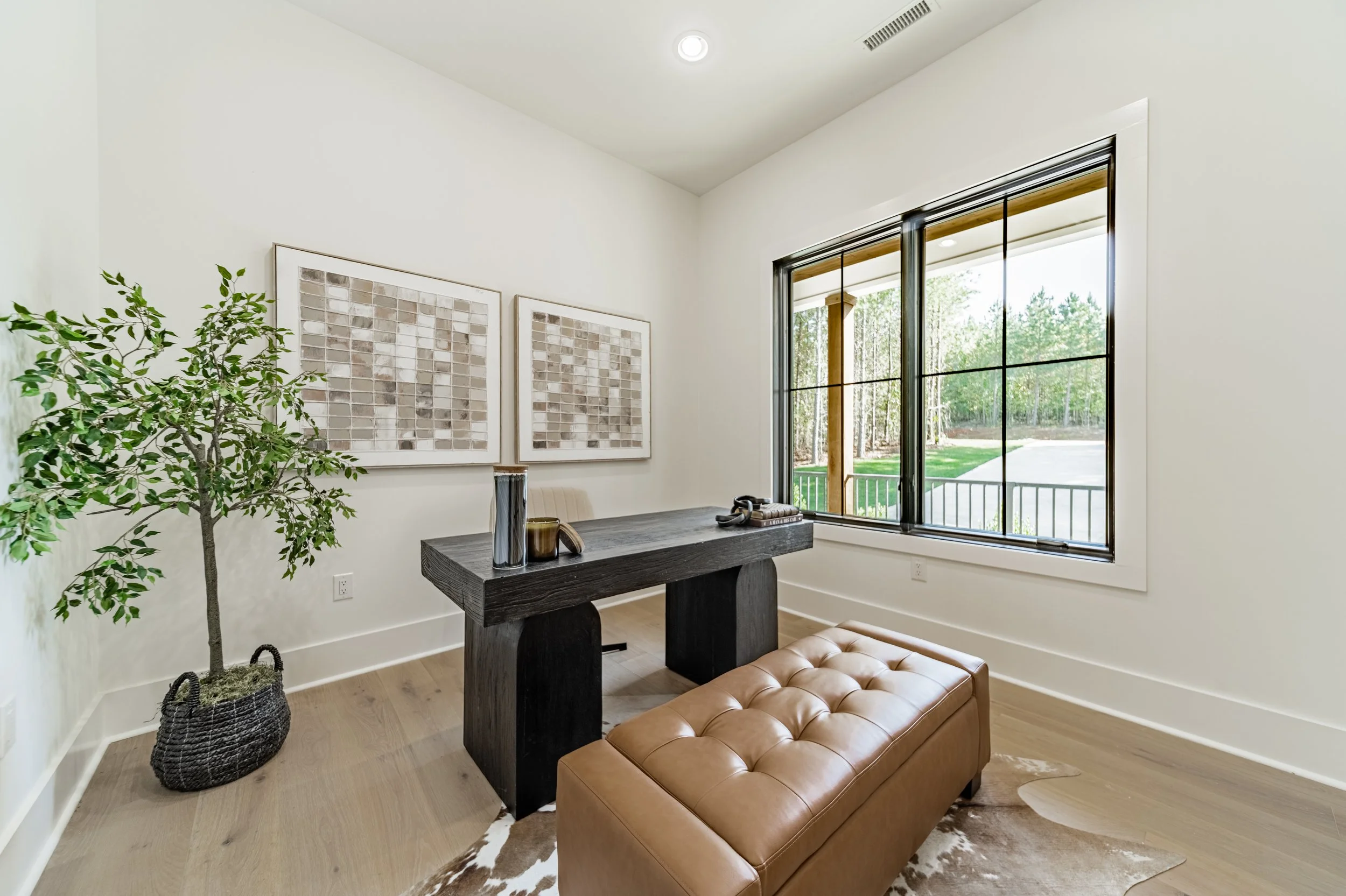 Interior room with large window, framed wall art, potted plant, brown leather ottoman, and black wooden desk.