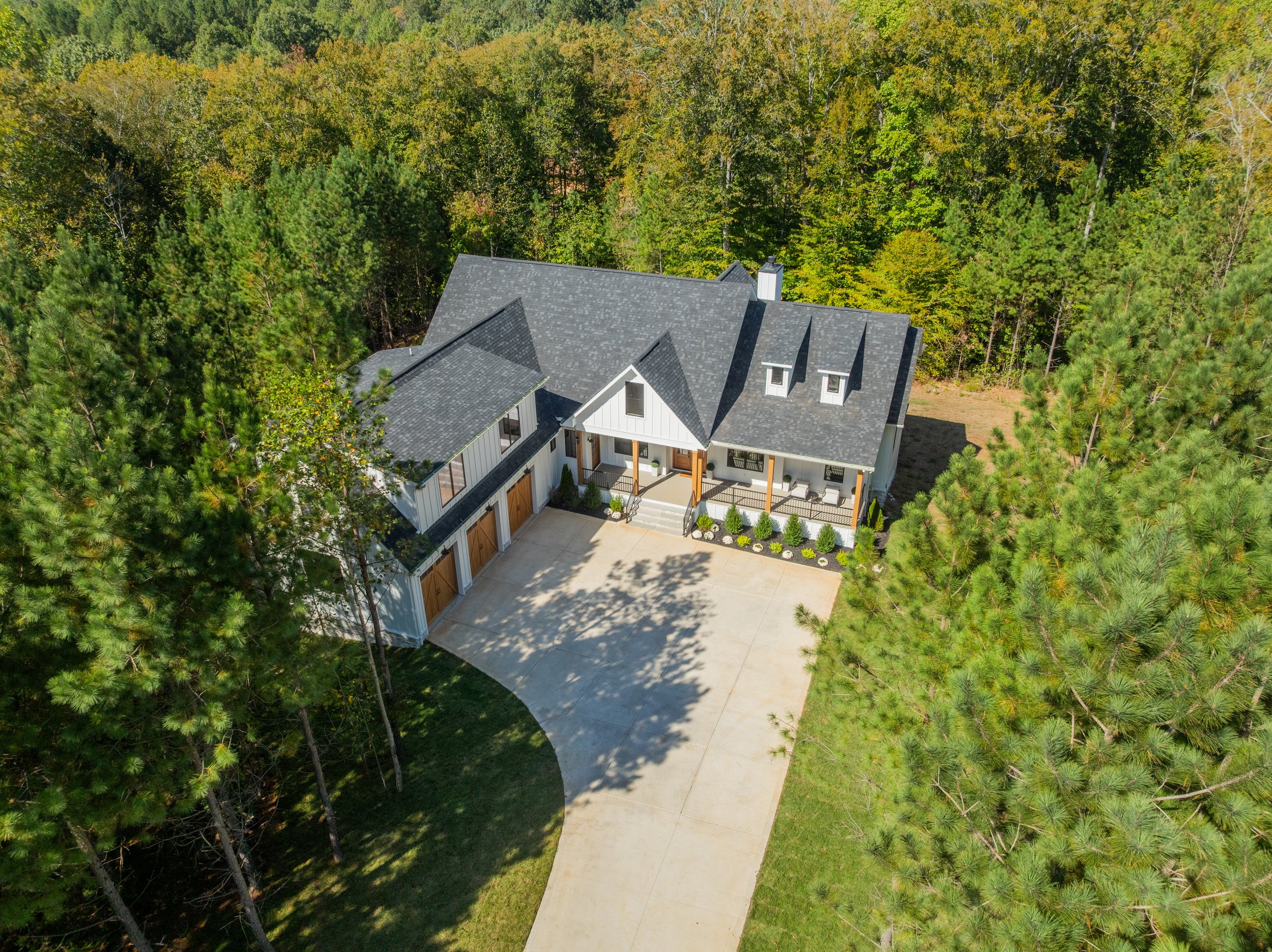A house surrounded by a dense forest of tall green trees, with a cement driveway leading up to the house.