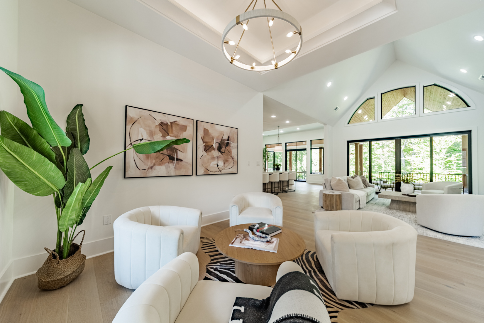 Bright, modern living room with large windows, white furniture, a circular chandelier, and art on the walls, with a view of a balcony and trees outside.