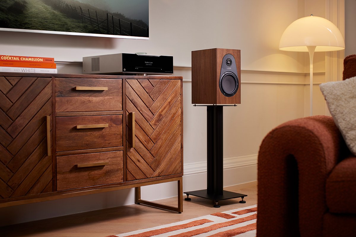 Stanza con mobile in legno, DSM Linn e diffusore acustico in legno su supporto nero, lampada da terra gialla, parte di un divano marrone con coperte beige e arancioni, e un televisore sopra il mobile.