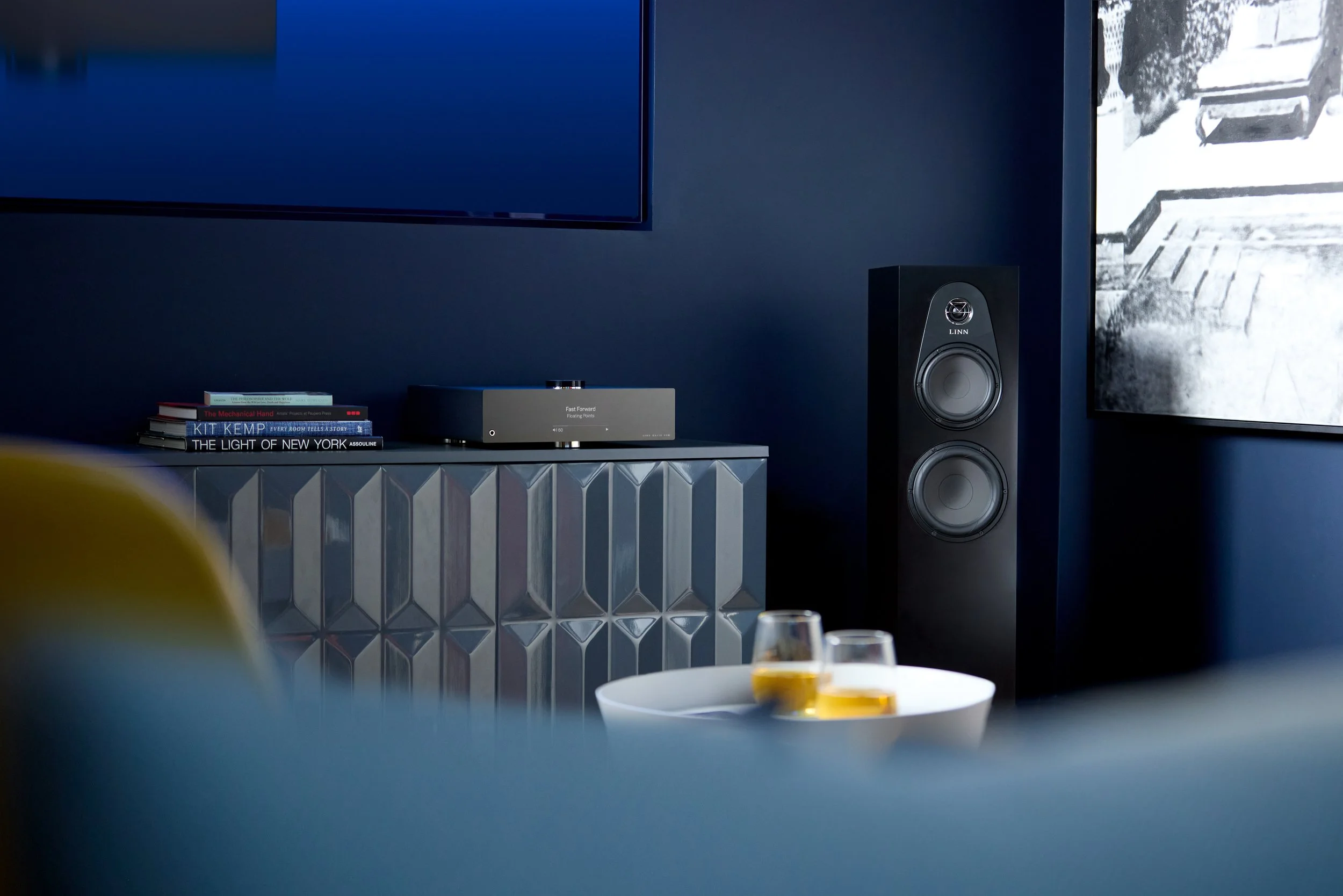 Un ambiente domestico moderno con un tavolo su cui ci sono alcune bevande, un sistema audio e una pila di libri. Sullo sfondo si vede una parete blu, un altoparlante grande e uno schermo TV.