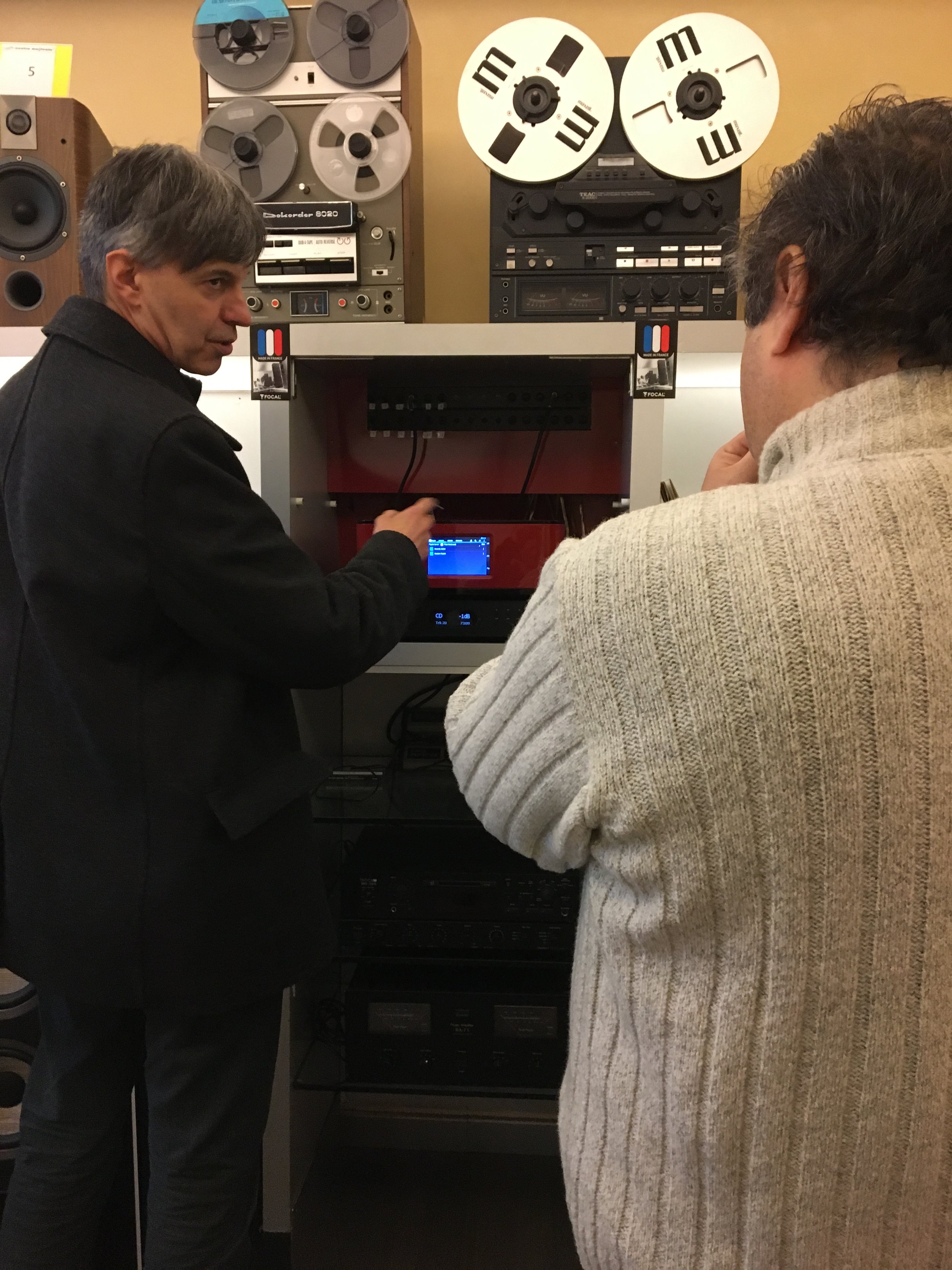 Due uomini con capelli castani e grigi, uno con giacca nera e l'altro con maglione grigio, stanno osservando correttamente un apparecchio audio con bobine di nastro e apparecchiature audio vintage.