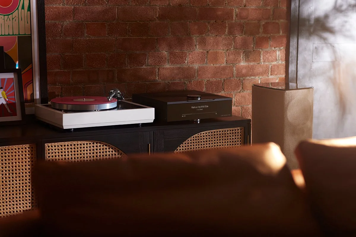 Setup audio con disco vinile e amplificatore su mobile con dettagli di arredamento in stile moderno, pareti in mattoni e tende beige.