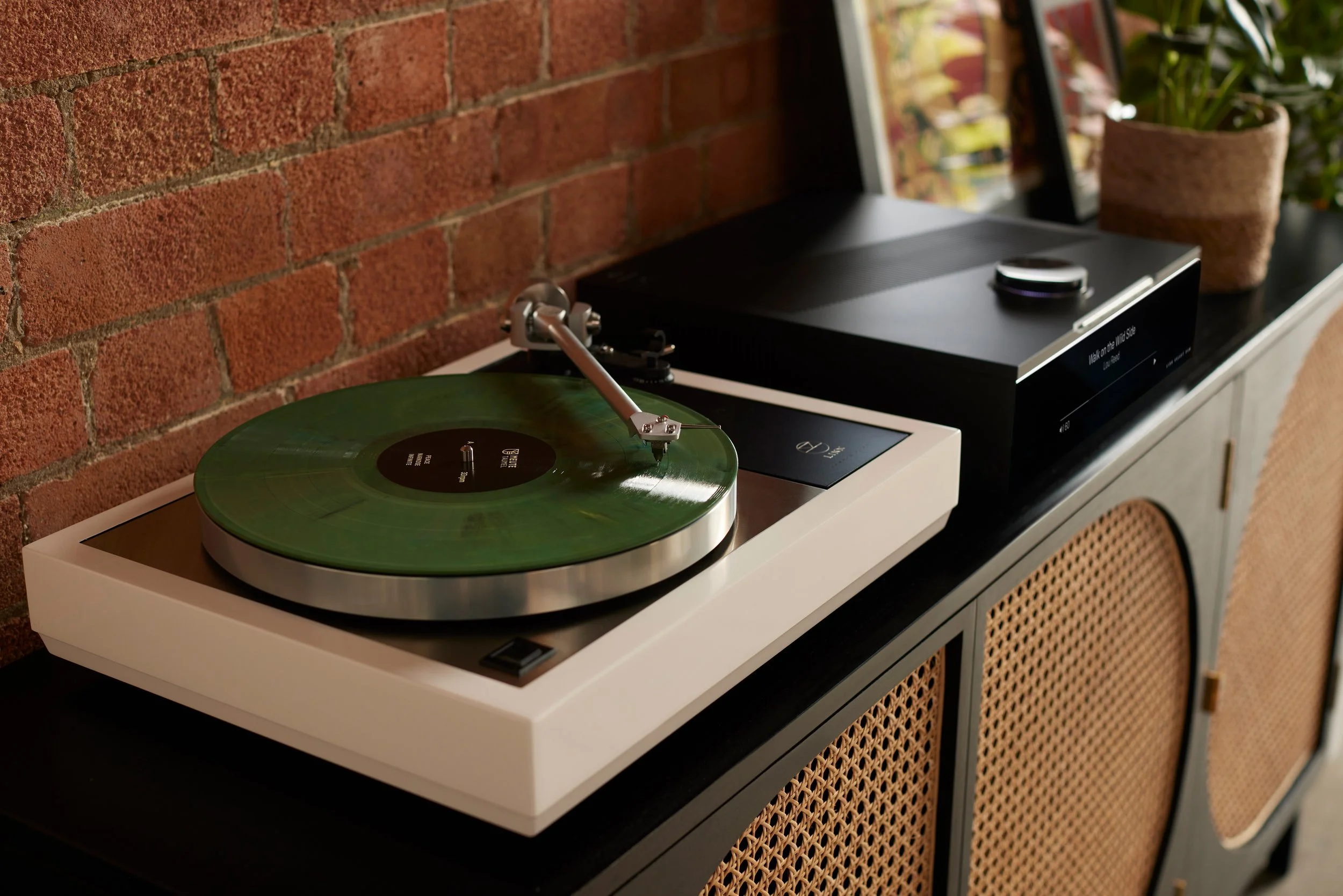 giradischi Linn Sondek LP12 con base bianca e vinile di color verde acceso, affiancata da un Linn DSM Sondek con display digitale, in un ambiente con muro di mattoni rossi e una pianta in un vaso di argilla.