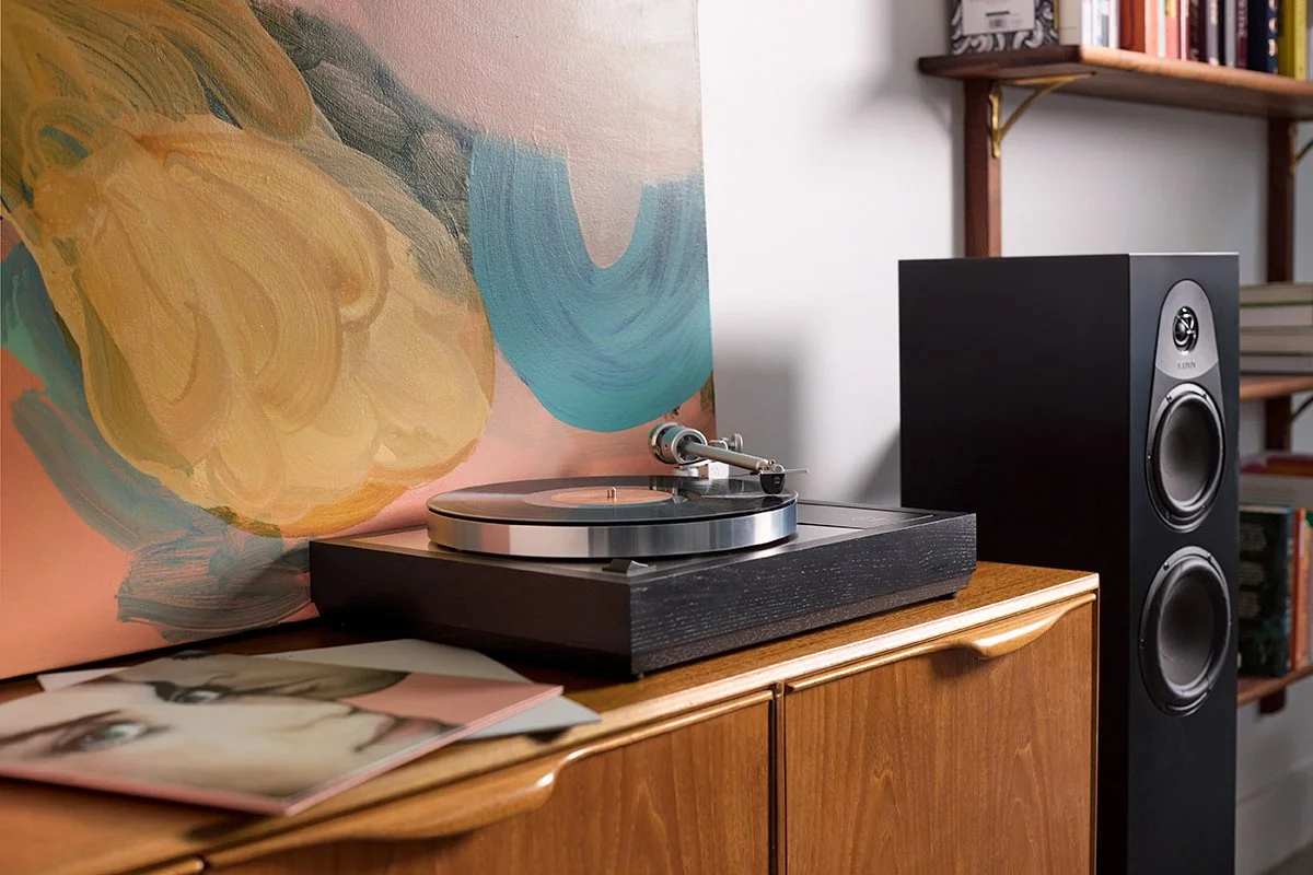 Un giradischi Linn Sondek LP12 nero su un mobile in legno con una grande opera d'arte astratta sullo sfondo e una libreria con libri a destra, accompagnato da due grandi casse acustiche nere.