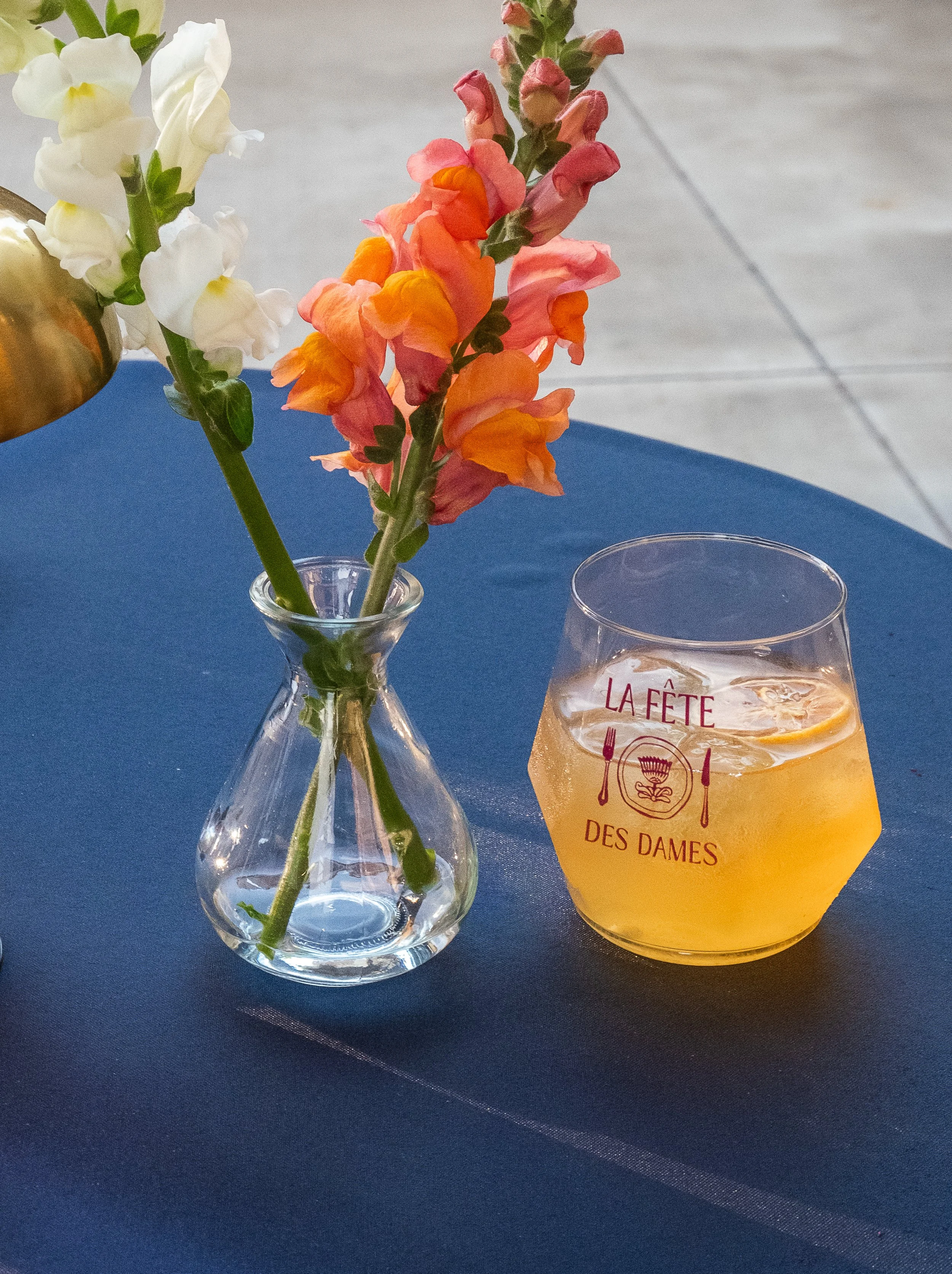 A tabletop decorative vase with flowers in it next to a wine glass with representing Le Fete De Dames a nashville event