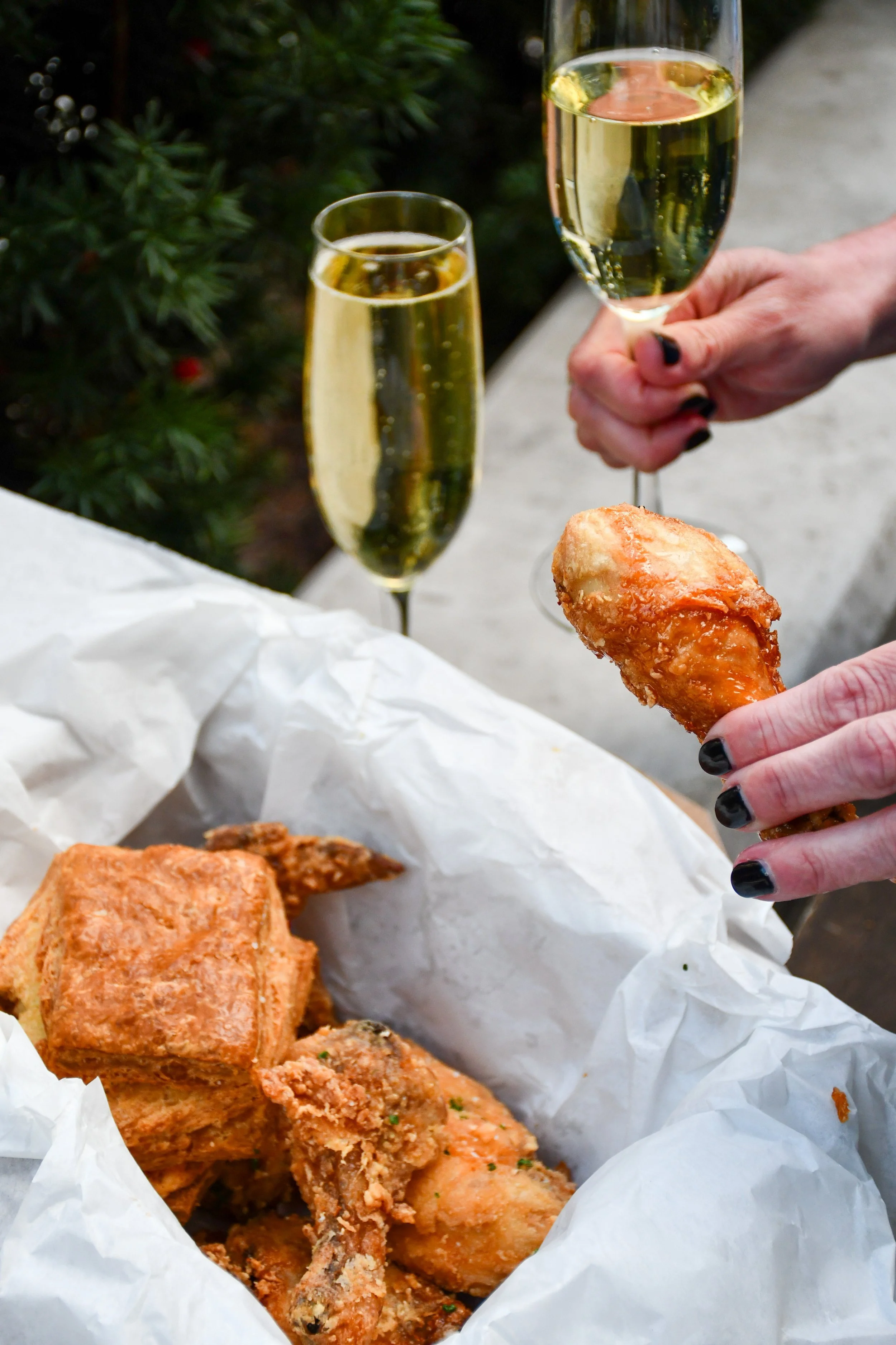 a basket of fried chicken, biscuits with a hand holding a piece of chicken and a glass of champagne in the other