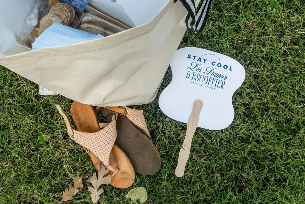A top-down view of a tote bag with food in it, a pair of sandals and a paper fan that says "stay cool les dames d'escoffier nashville"