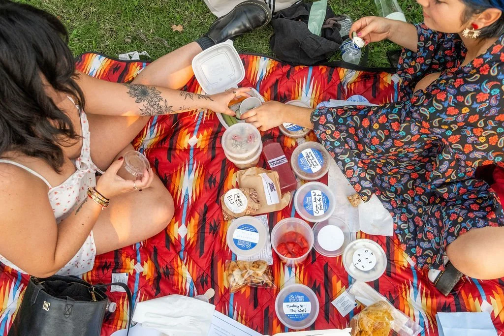  Two women sitting on a picnic blanket sharing small containers of food while holding drinks at Les Dames Nashville’s picnic event at Centennial park 