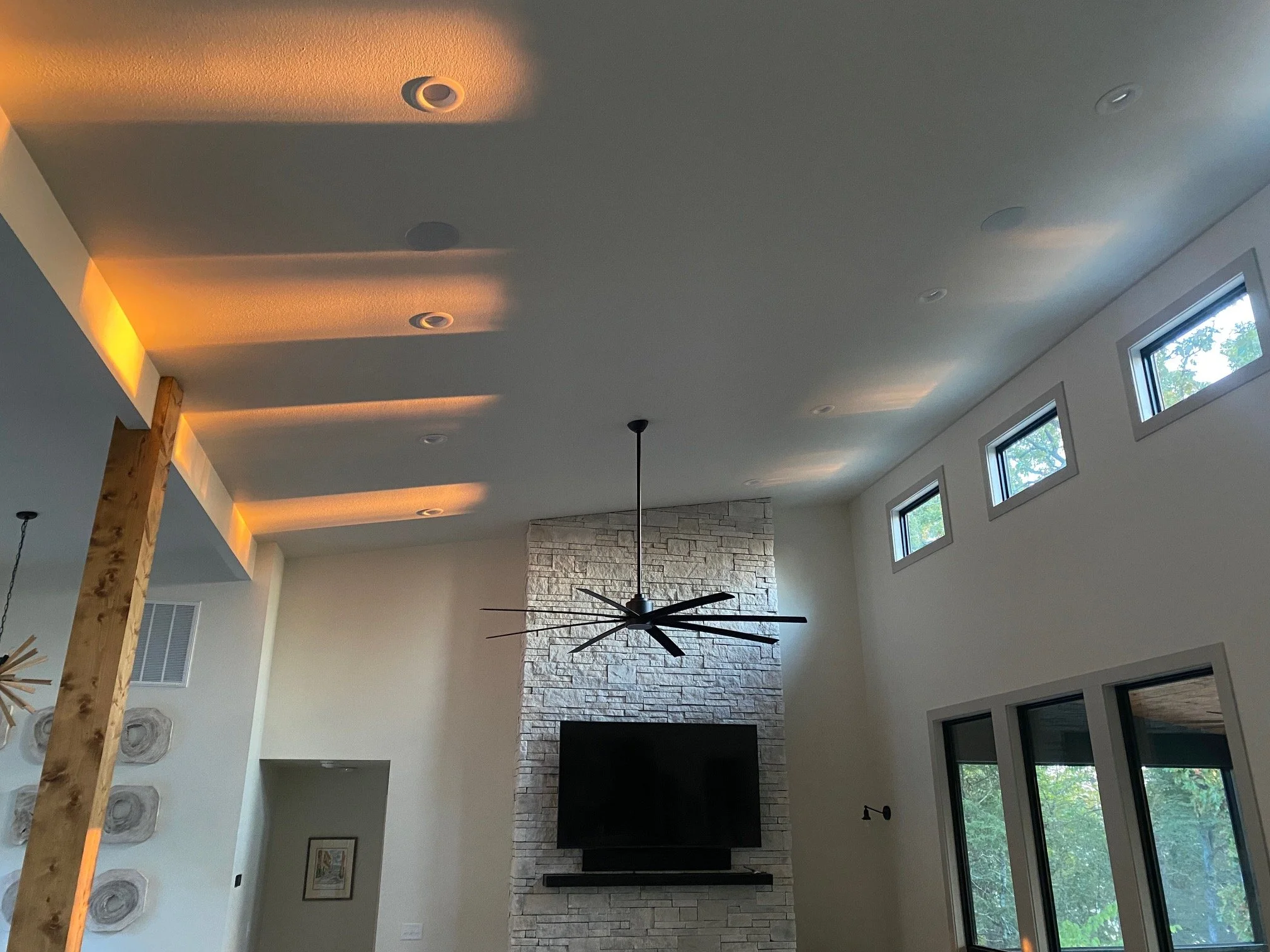 A modern living room with a ceiling fan, stone fireplace, and large windows, illuminated by natural light.