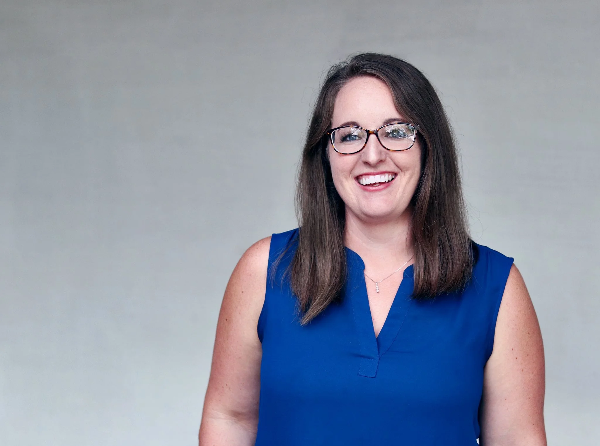 A woman with long brown hair, glasses, and a bright smile, wearing a sleeveless blue top, standing against a plain gray background.
