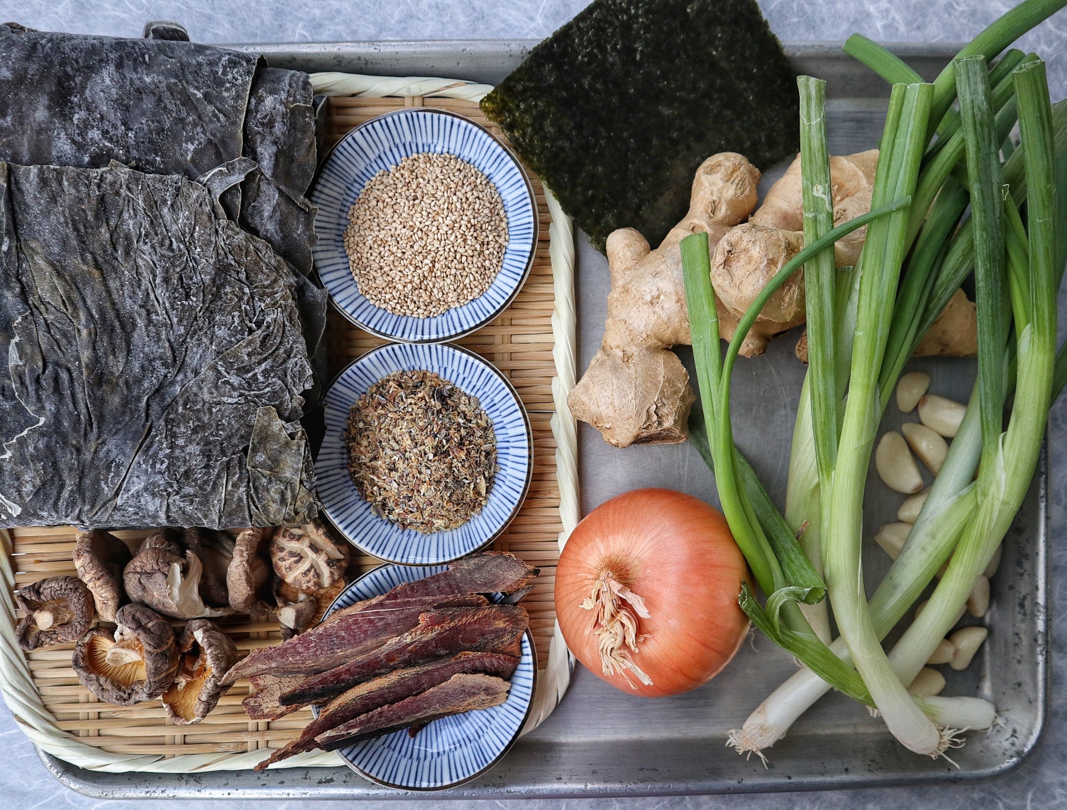 Assorted Japanese ramen ingredients on a bamboo tray — dried nori seaweed, sesame seeds, fresh ginger root, dried ginger, green onions, garlic, mushrooms, chili peppers, and onion, used at Satori Ramen Bar in Columbus, Ohio.