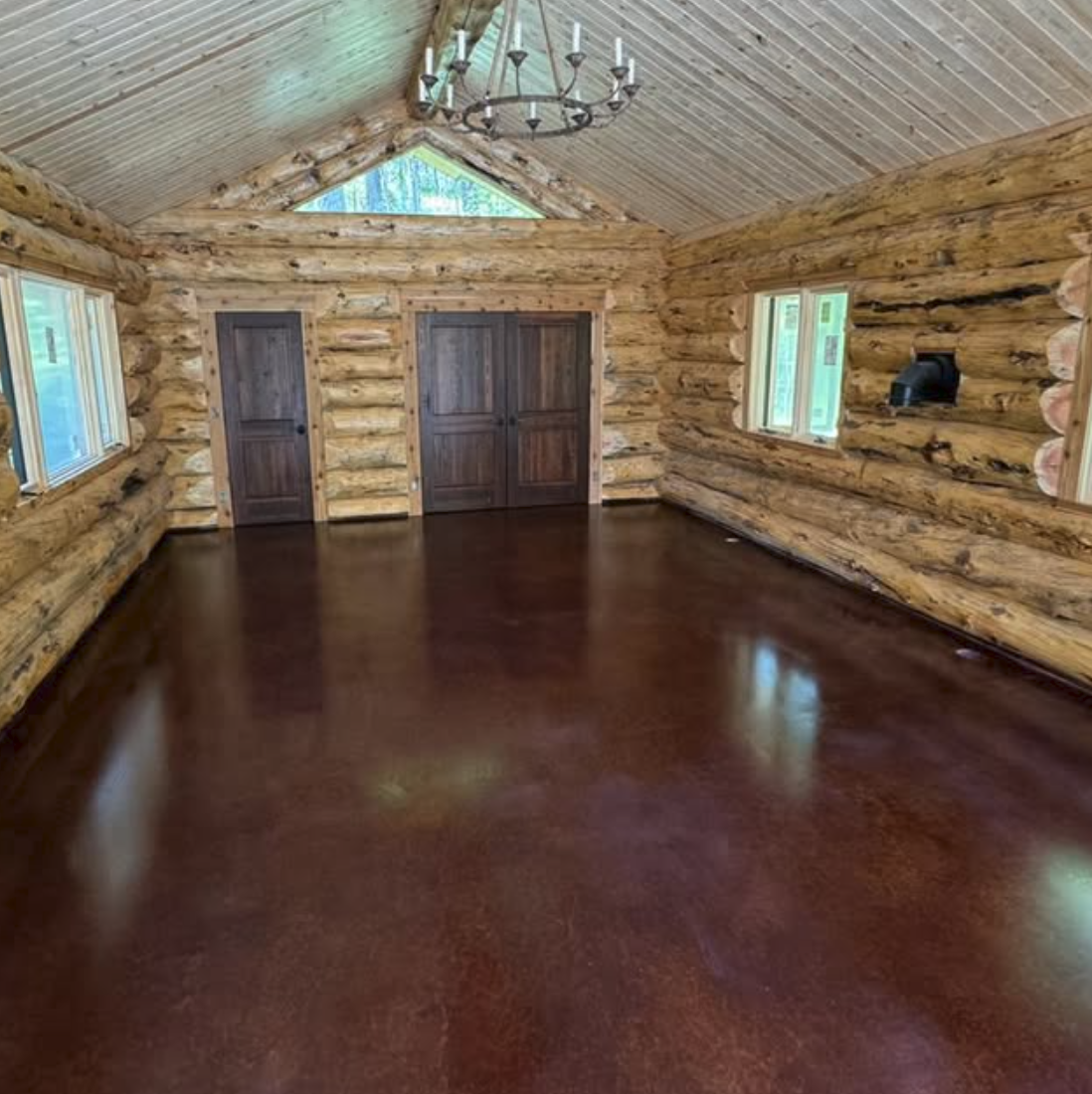 Empty log cabin-style room with wooden log walls, wooden ceiling, dark hardwood floor, chandelier, and several windows.