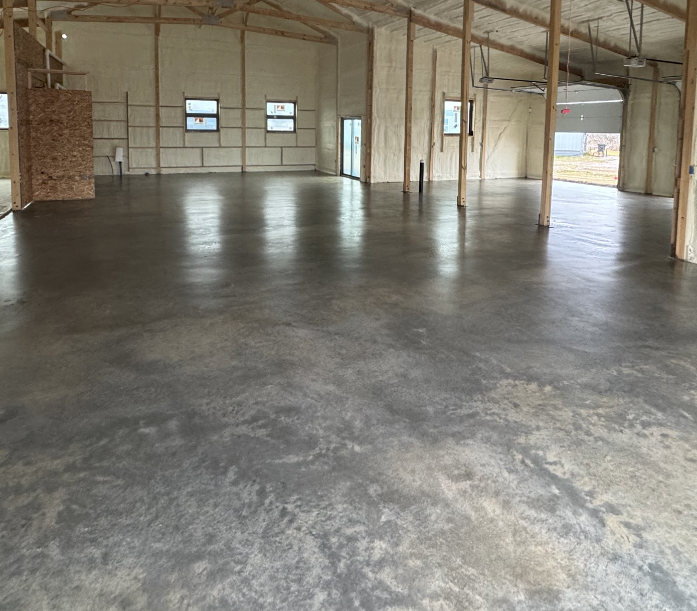Empty garage with polished concrete floor, unfinished walls, exposed wooden beams, and a partially open garage door