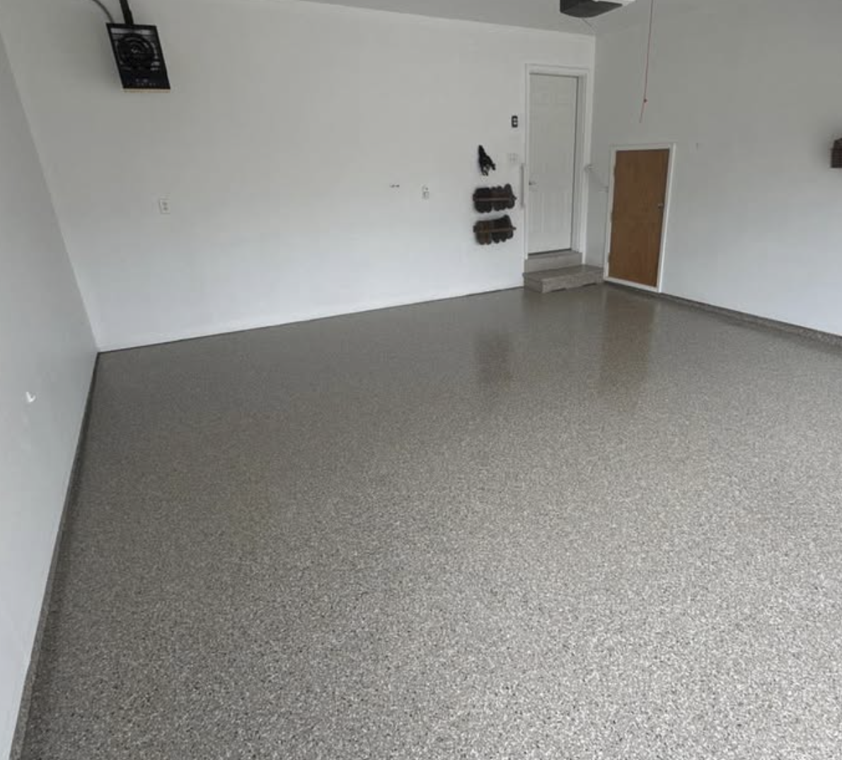 Empty garage with speckled gray flooring, white walls, and a white door with steps leading up in the background. There are wall-mounted shoe racks and a small wooden door.