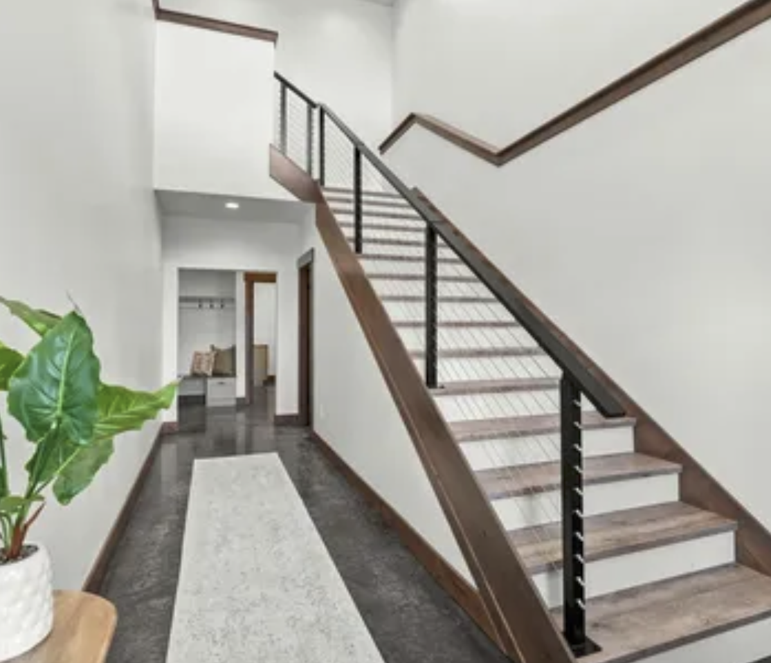 Interior of a modern home hallway featuring a staircase with wooden steps, black metal railing, white walls, a light-colored rug on a dark floor, and a potted green plant.