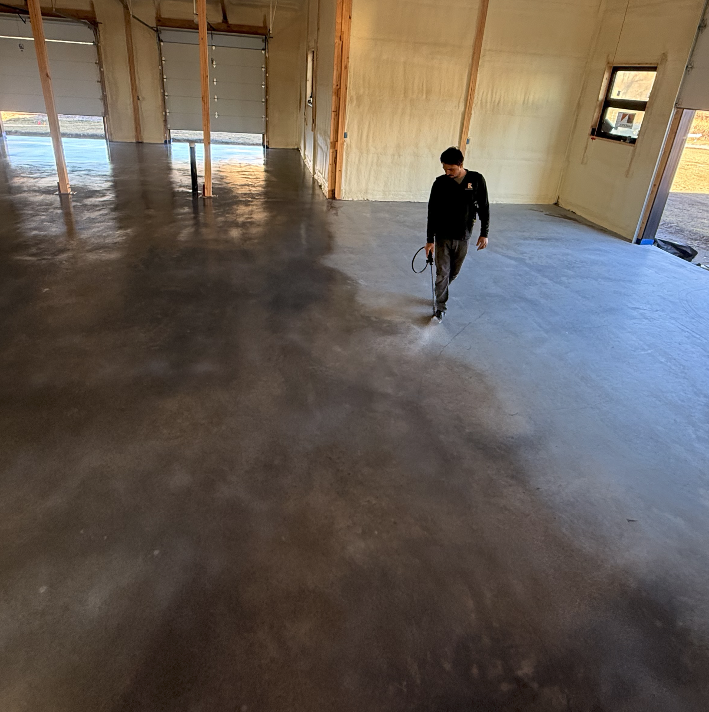 A person walking inside a newly constructed garage with a polished concrete floor. The garage has unfinished walls, several large garage doors, and a window on the right side. The person appears to be inspecting or working on the floor.